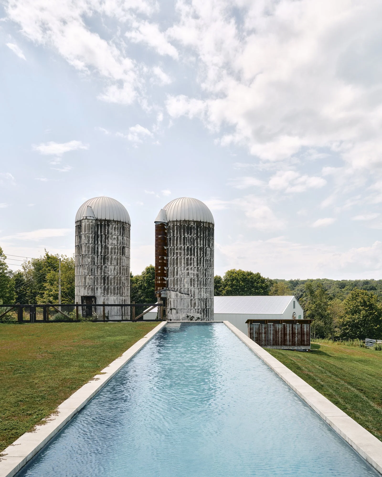 A narrow rectangular water feature outdoors leading to two large, weathered silos with domed tops and a rural farm building in the background, under a partly cloudy sky.