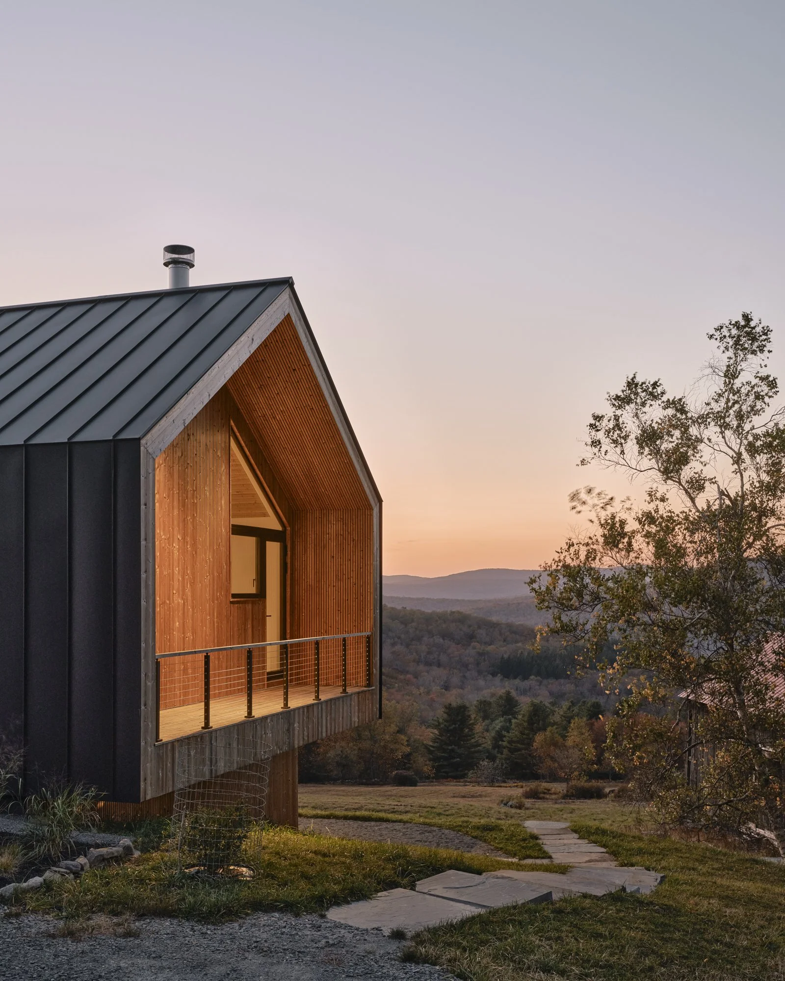 Modern house with metal roof and wooden porch, overlooking a scenic landscape with trees and hills at sunset.