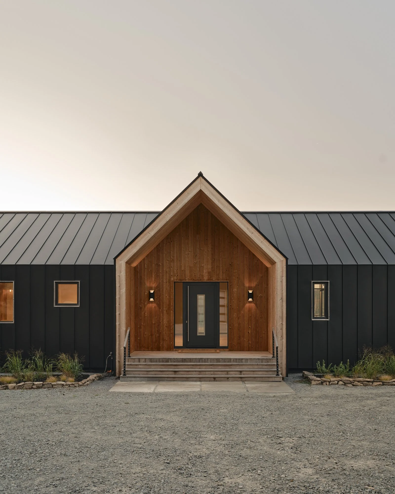 Modern house with a gabled wooden entrance, black metal siding, and small windows, set on a gravel lot with landscaping.