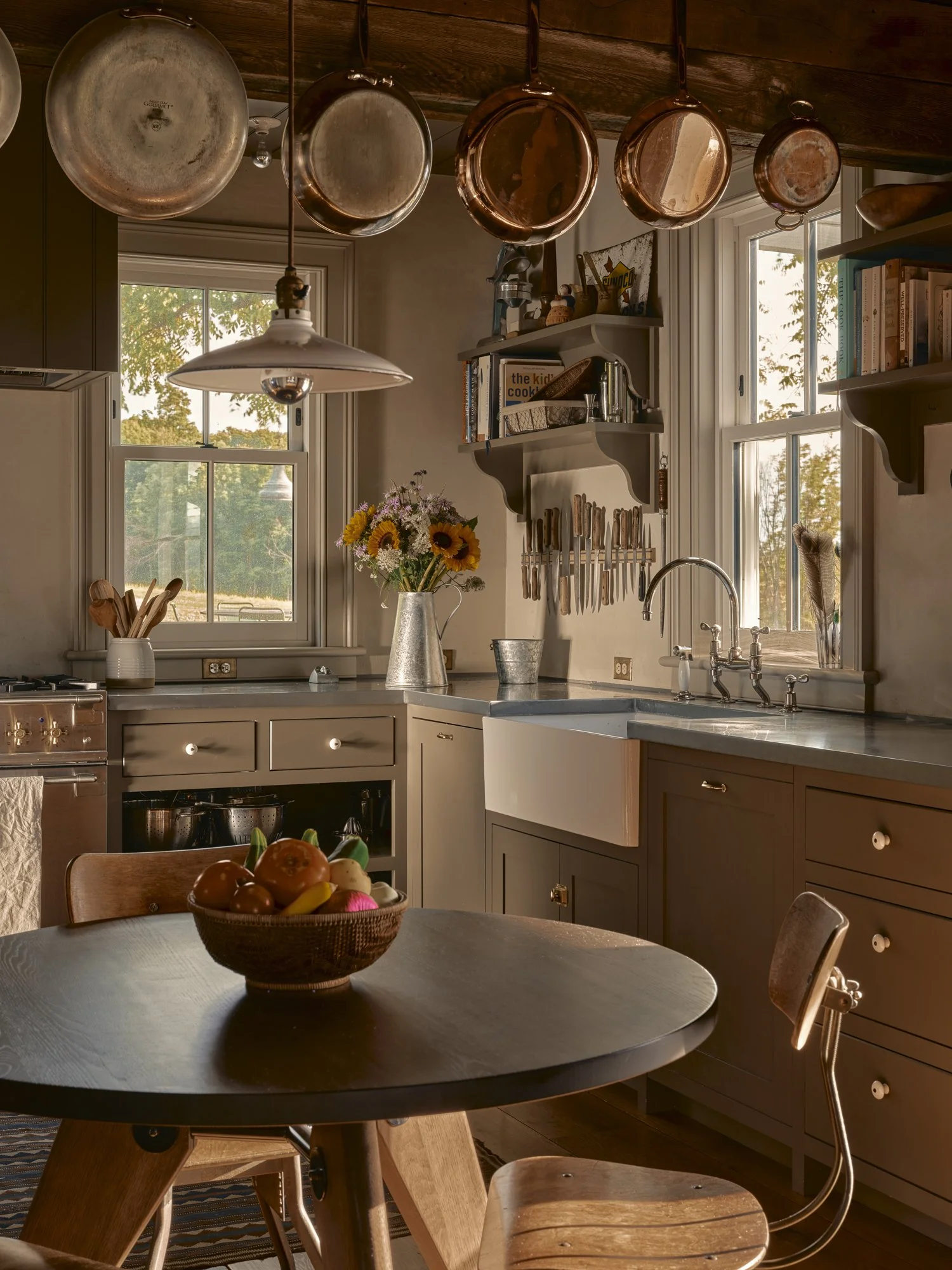 Cozy kitchen with hanging copper pots, white and gray cabinetry, a round wooden table with a basket of decorative fruits, and large windows letting in warm natural light.