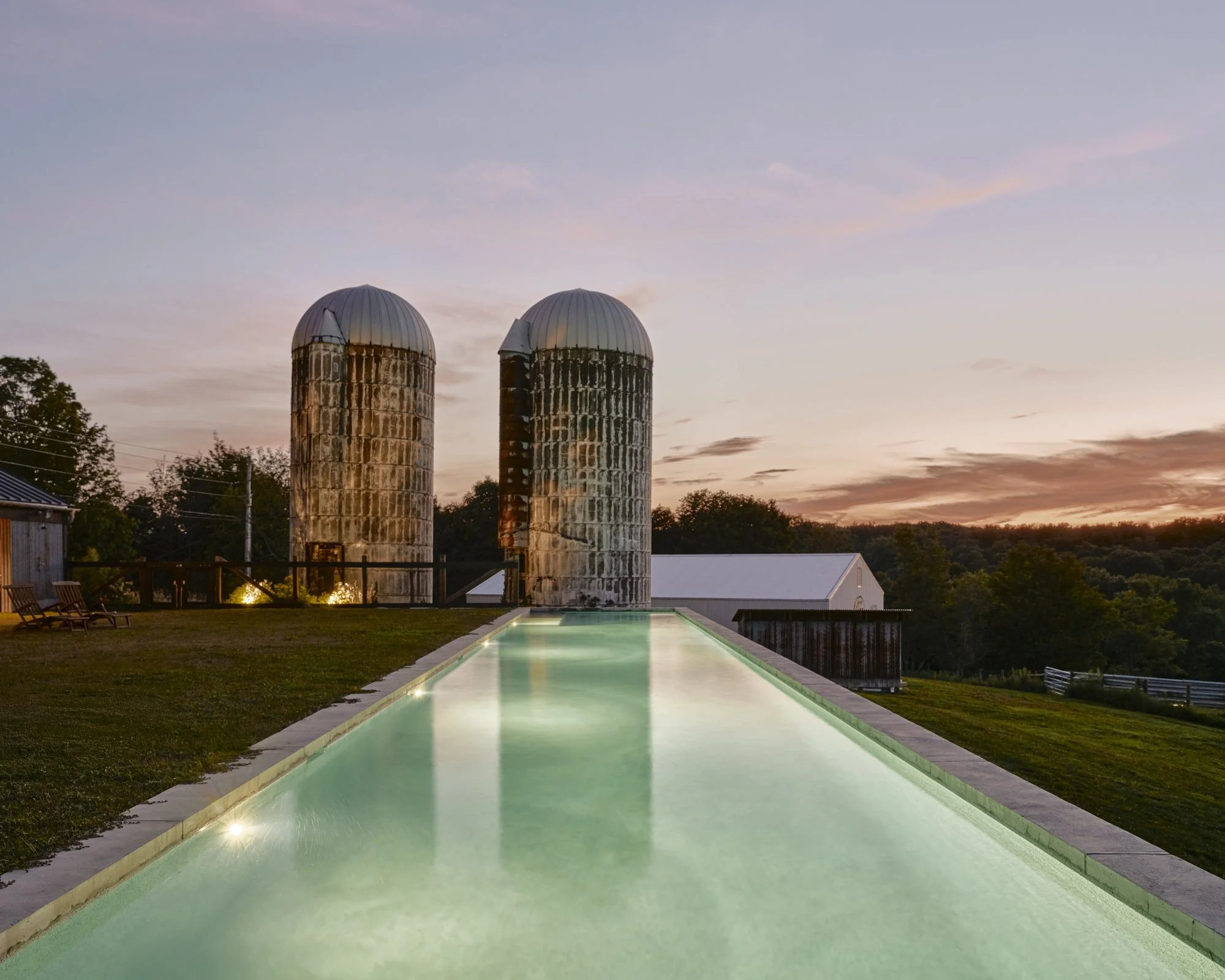 A swimming pool at sunset with two silos and a white barn in the background.
