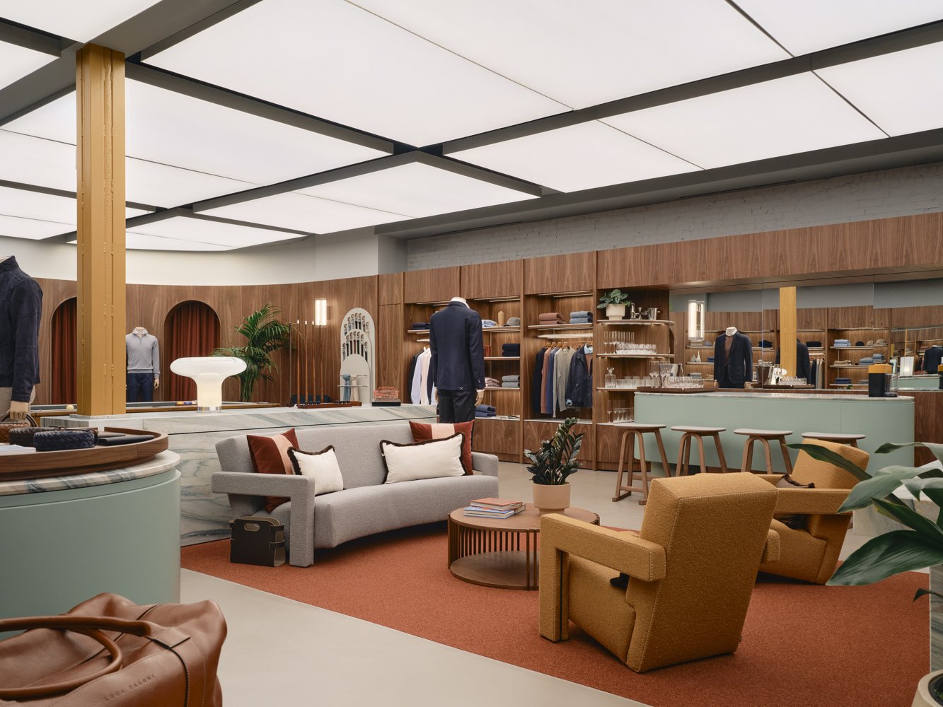 Stylish clothing boutique with seating area, wooden shelves displaying folded clothes and glassware, mannequins dressed in suits, and modern decor with plants and warm lighting.