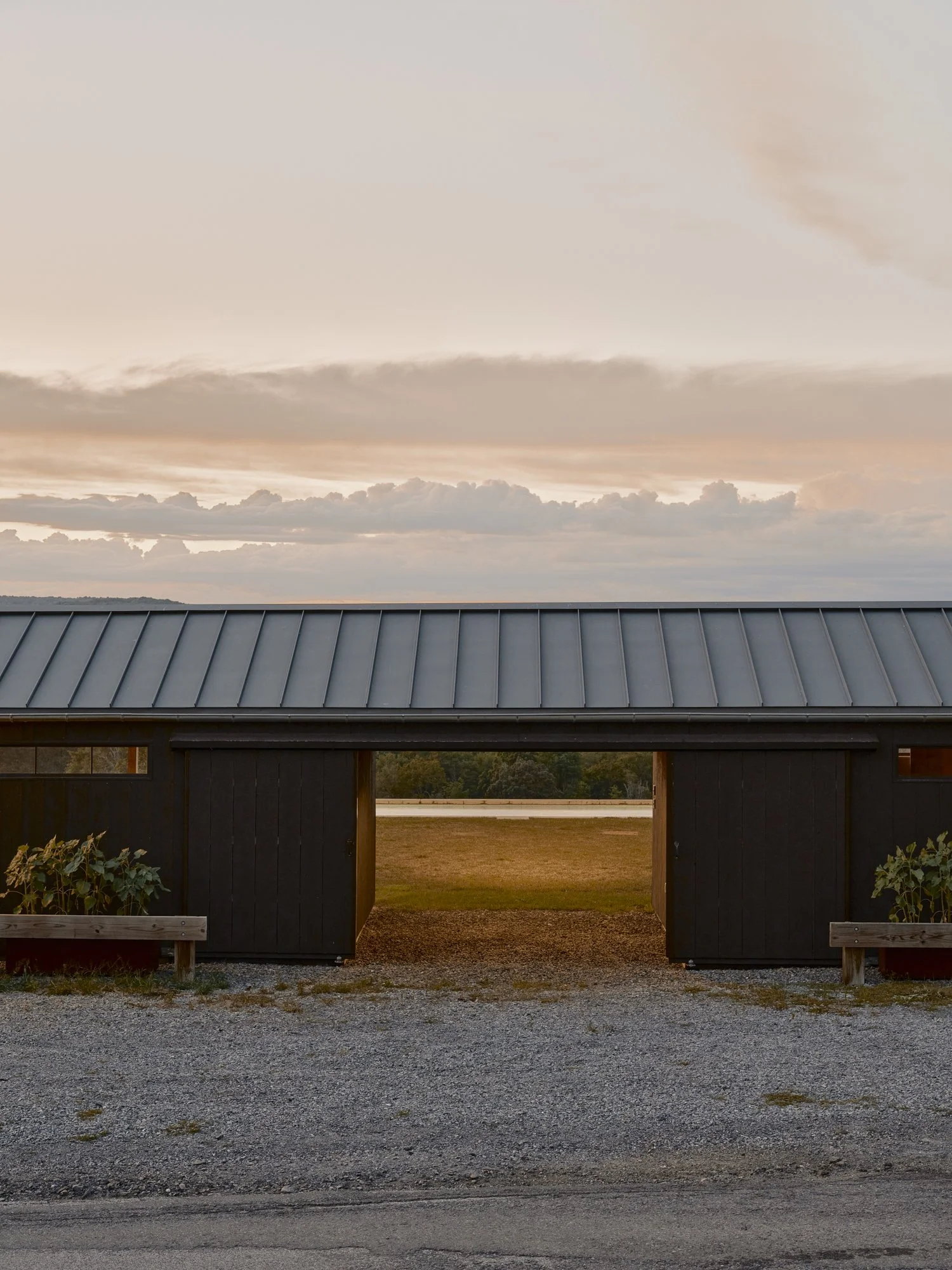 A modern dark-colored building with a metal roof and an open entrance in the center, flanked by two small flower beds on each side, with a distant landscape and a cloudy sky at sunset or sunrise.