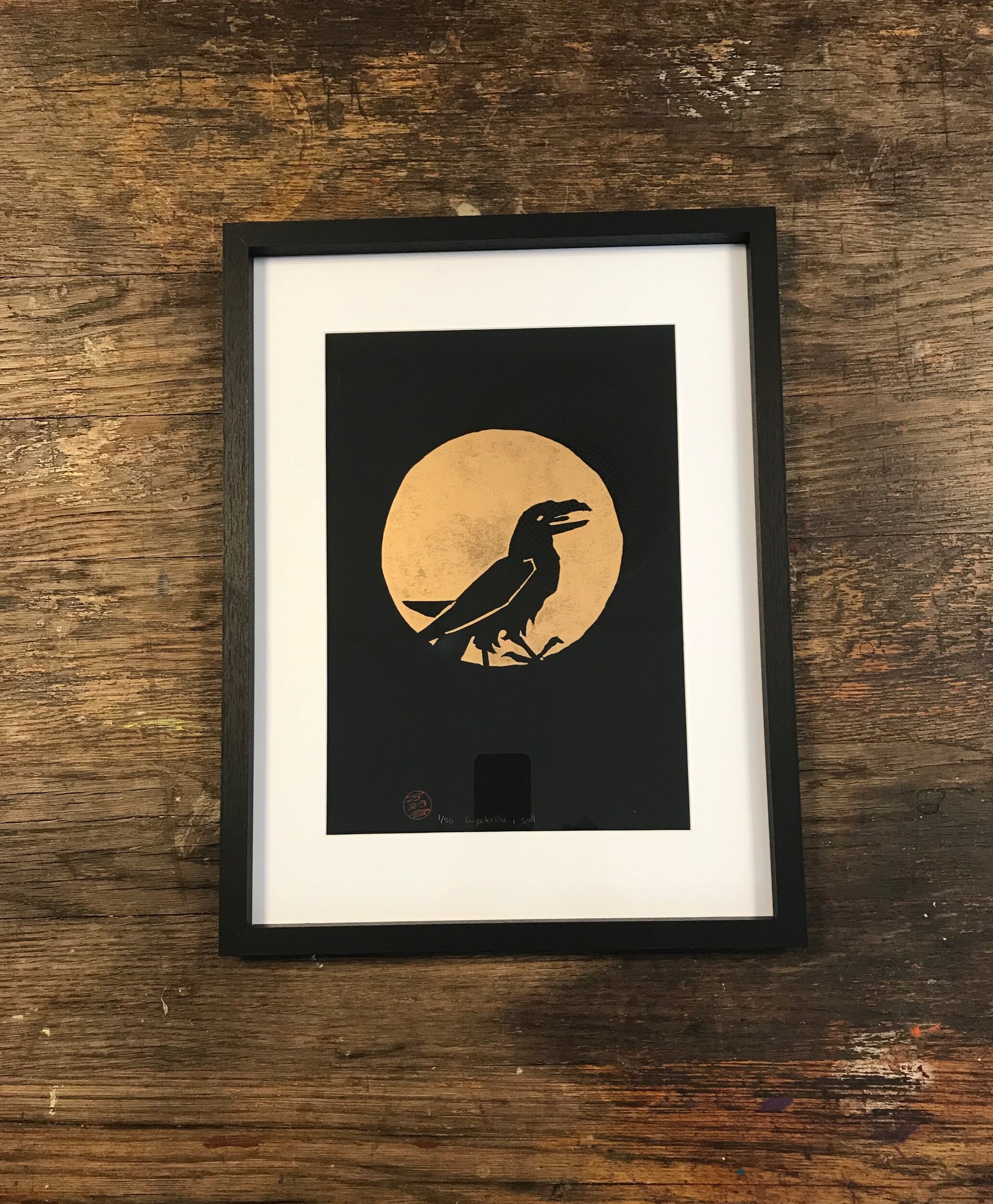Kråka i sentrum trykket i gull på sort kunstpapir, innrammet