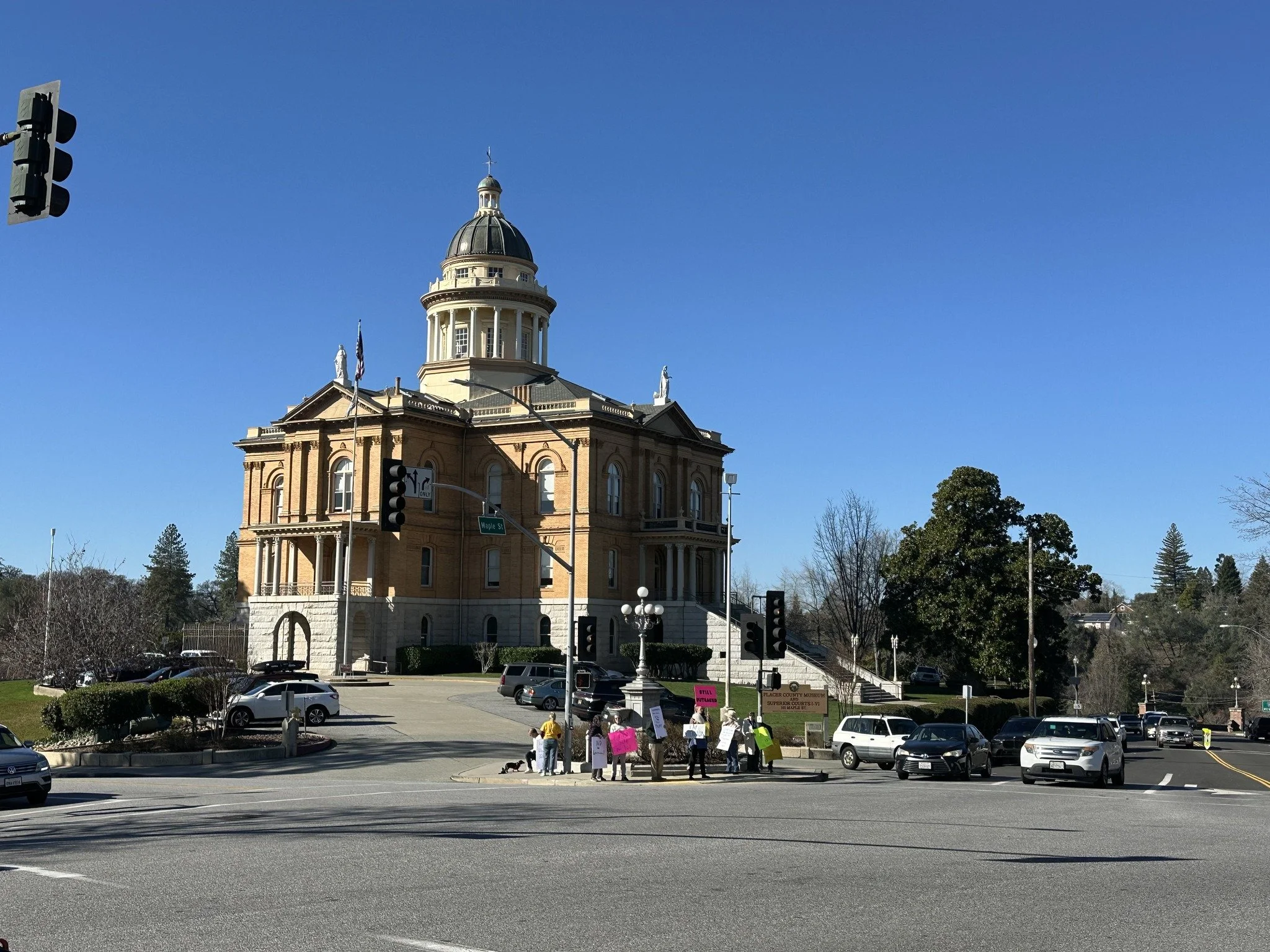 A historic building with a large dome and columns, situated on a busy street corner with cars and people gathered nearby, some holding signs.