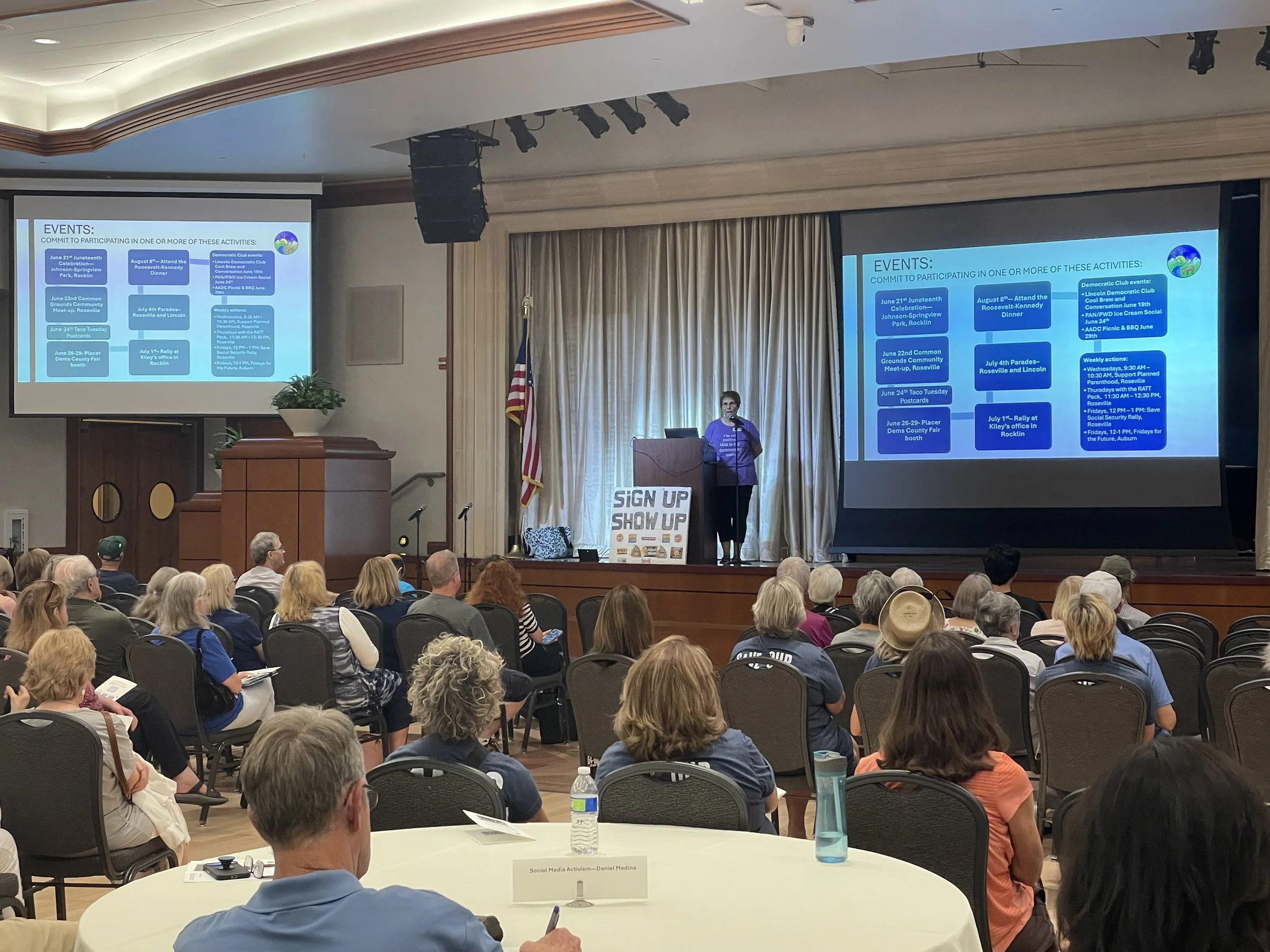 A large conference room filled with people listening to a speaker standing on stage. The stage has a large screen displaying event information. The audience is seated at round tables with name tags, water bottles, and papers. The room has beige curta