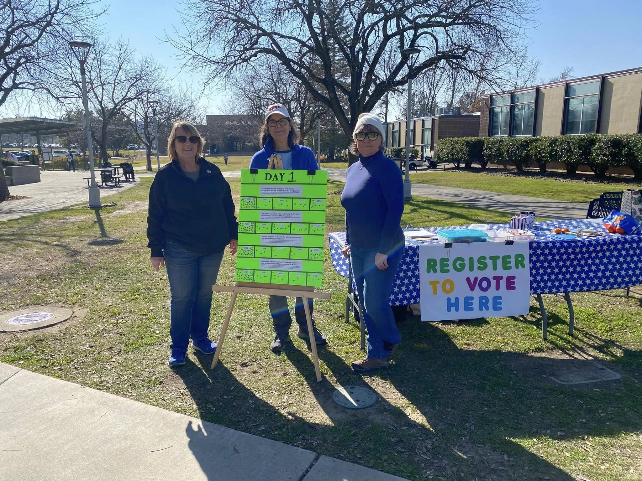 Three women standing outdoors at a voting registration table on a sunny day, with leafless trees and a building in the background.