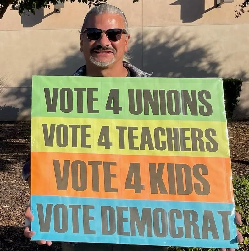 Person holding a sign with colorful stripes promoting voting for unions, teachers, kids, and Democrat, wearing sunglasses outdoors.