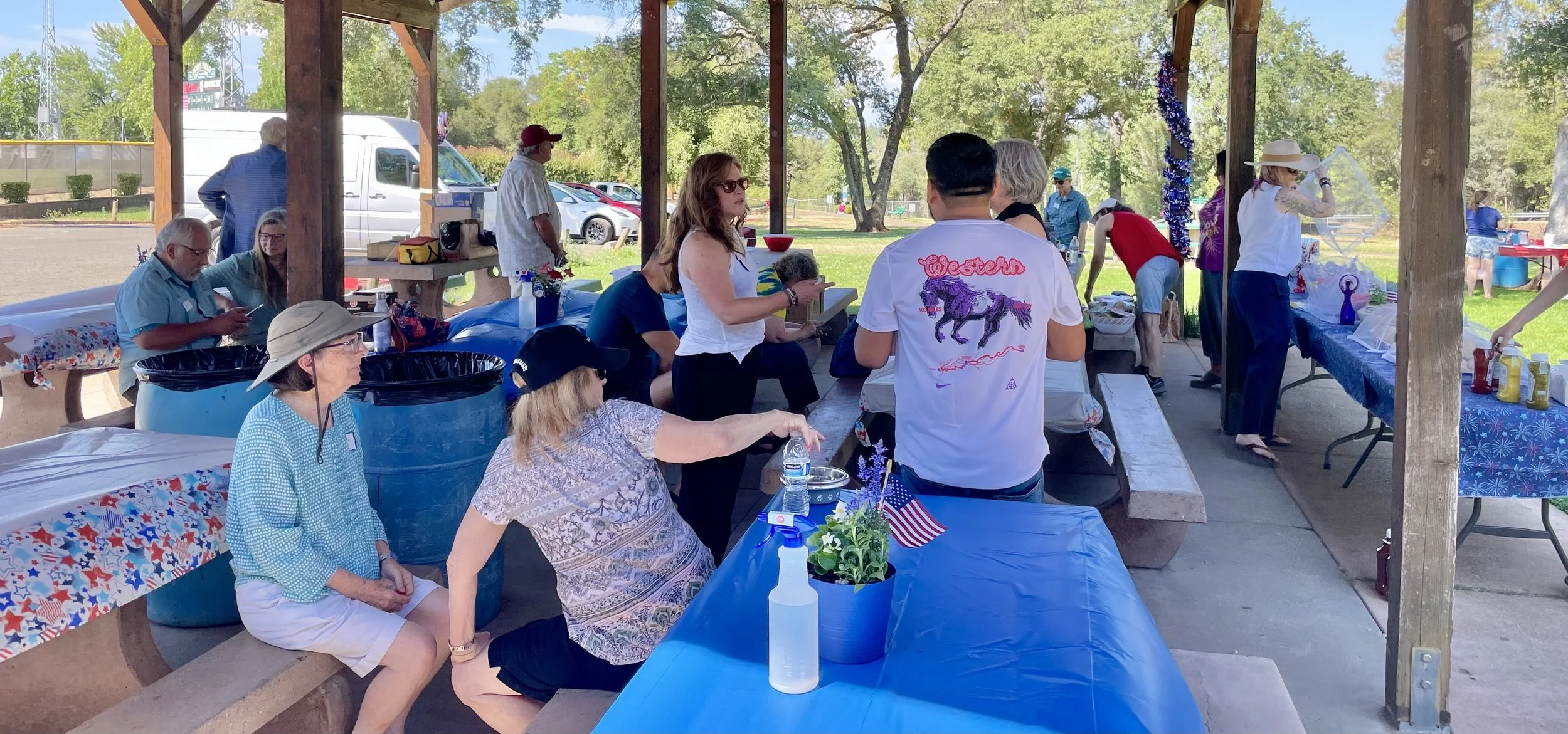 People gathered at a park pavilion during a patriotic outdoor event, with tables decorated in red, white, and blue, and some wearing casual summer clothes