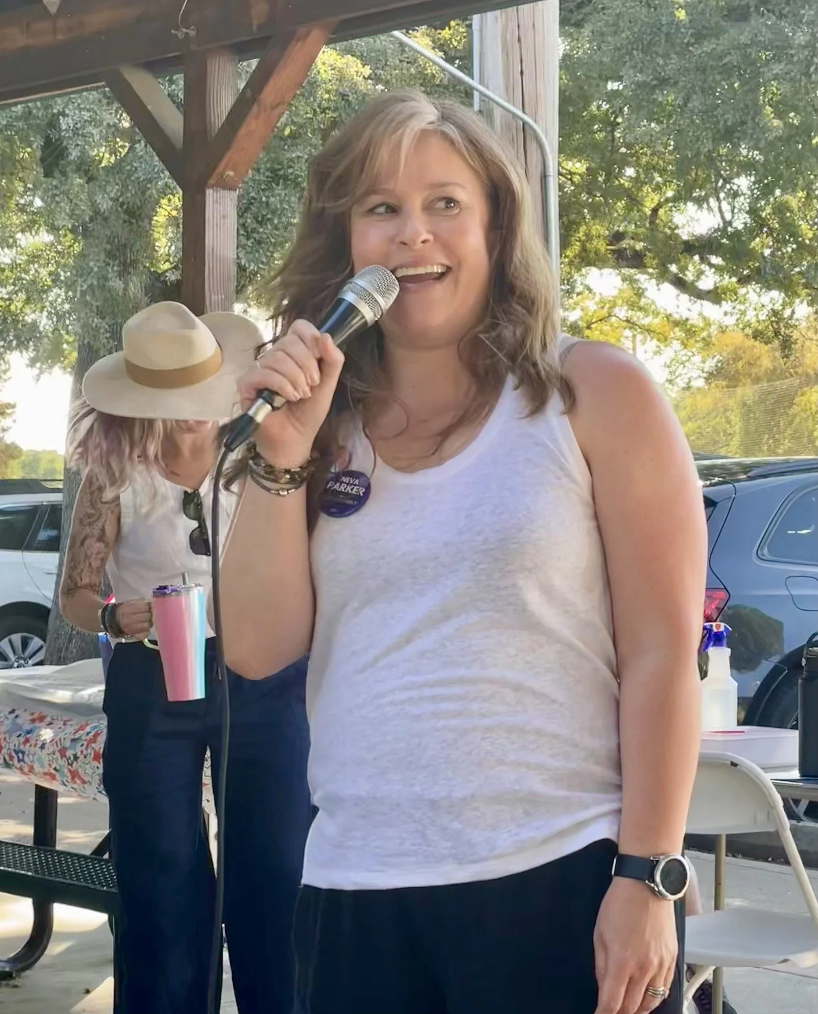 A woman with brown, shoulder-length hair, wearing a white tank top and a smartwatch, holding a microphone and smiling at an outdoor gathering with trees and parked cars in the background.