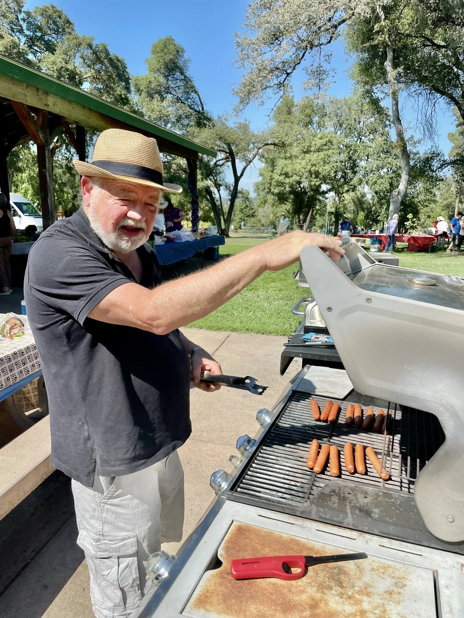 An elderly man with a beard, wearing a straw hat, black shirt, and light-colored shorts, is grilling hot dogs on a barbecue outdoors in a park during daytime. He is holding tongs and smiling, with other people and picnic tables visible in the backgro