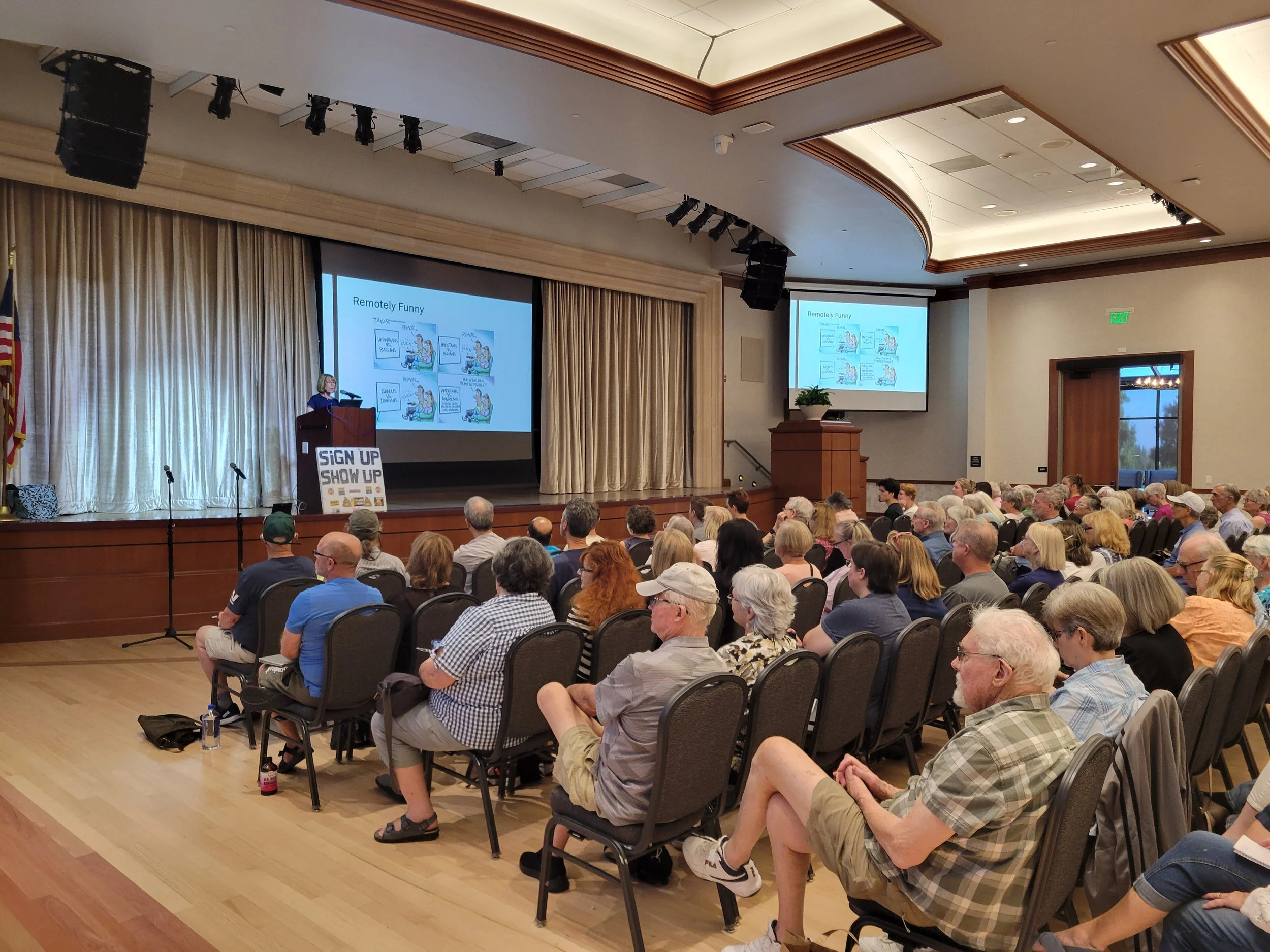 A large group of people seated in a conference room, watching a presentation on two big screens. The presenter stands at a podium on the stage, and a sign advertising sign-up and show-up is visible near the stage. The room has beige curtains, wooden 