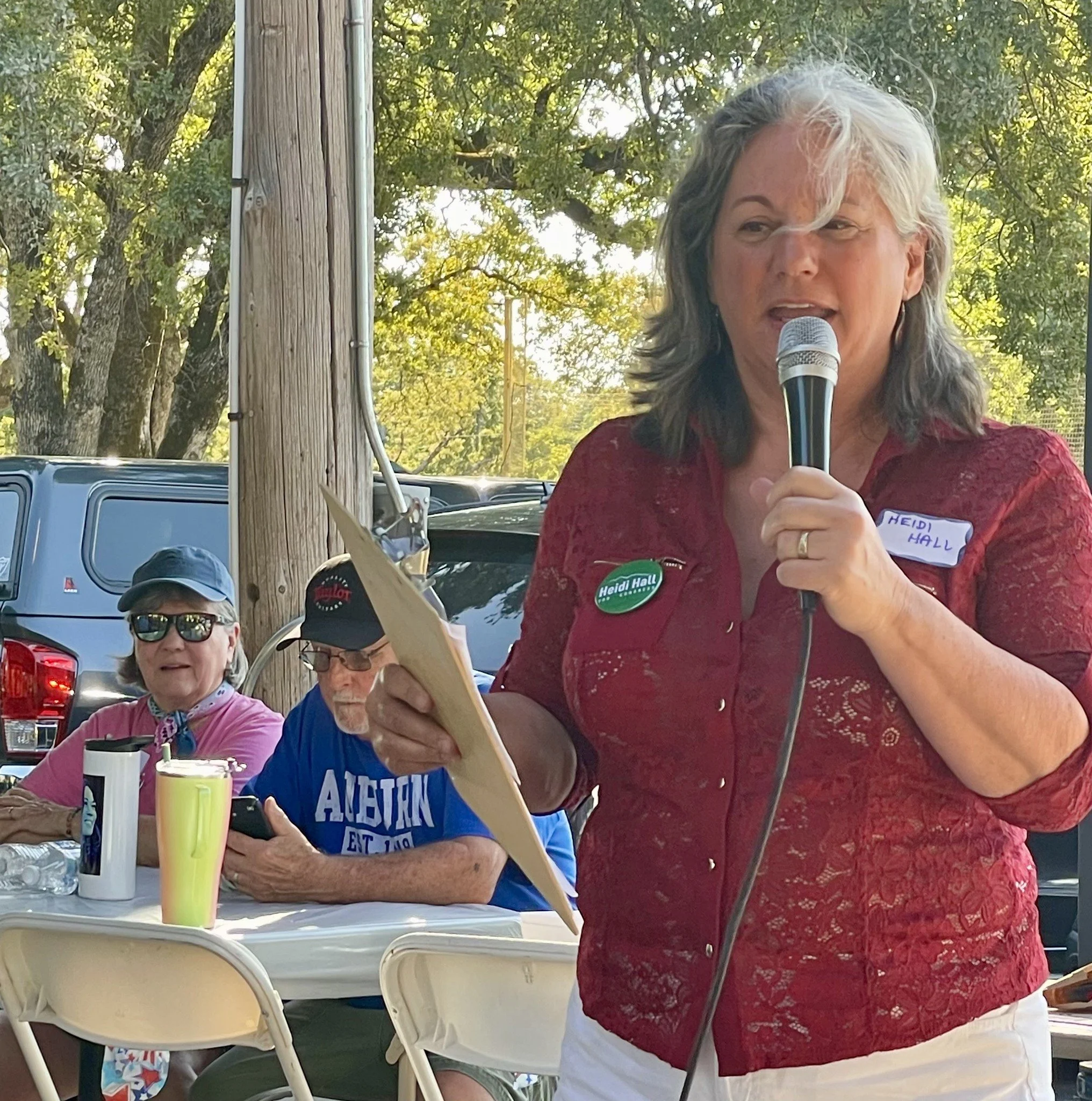 A woman with gray hair holding a microphone and reading from a paper at an outdoor event, wearing a red lace top and a green campaign button. Two people sitting at the table behind her, one with sunglasses and a hat, the other with glasses and a cap,