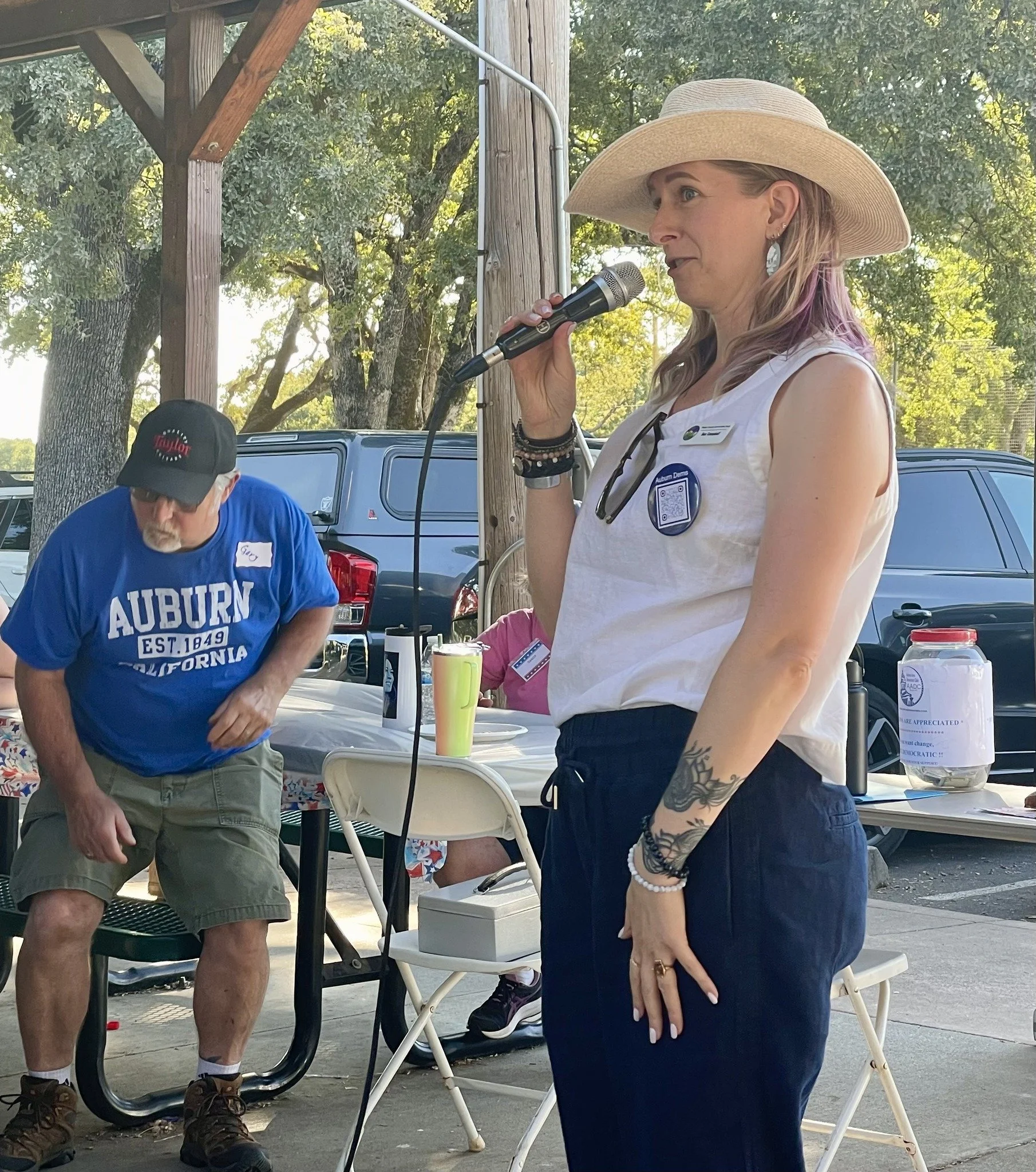 A woman with blonde hair, wearing a large sun hat, sleeveless white top, navy pants, and various bracelets, speaking into a microphone at what appears to be an outdoor gathering or event.