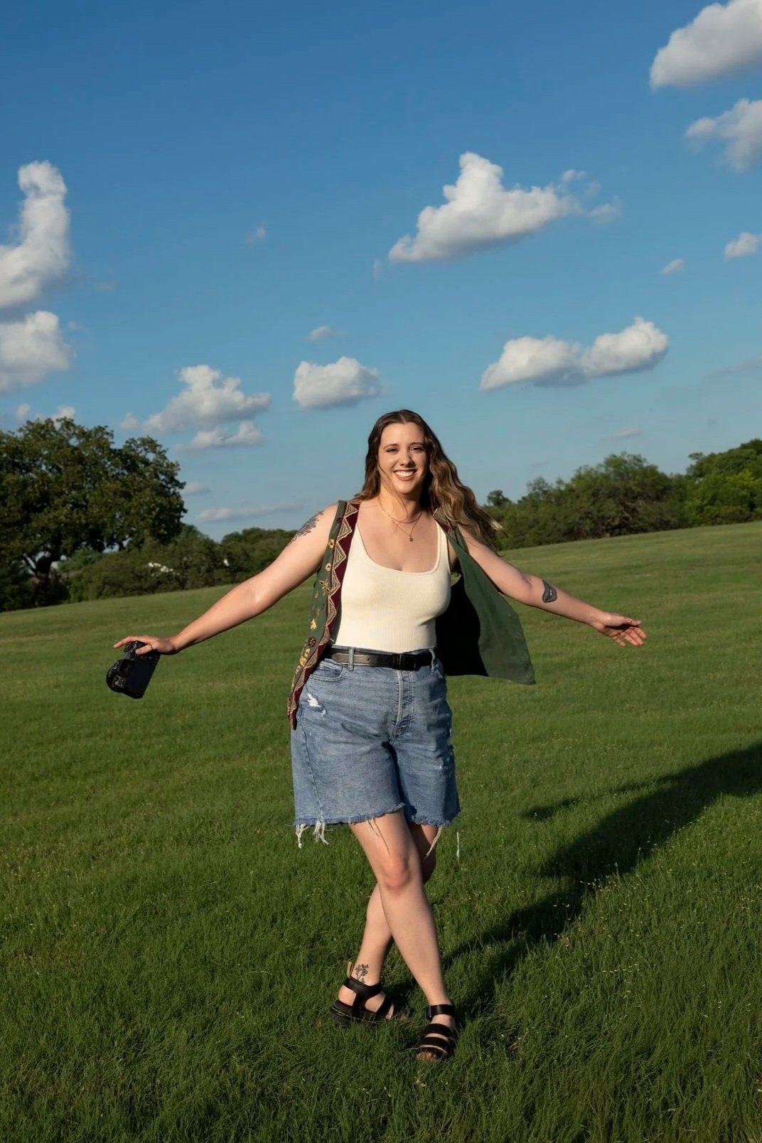 A smiling woman standing in a grassy field with trees in the background under a partly cloudy sky, holding a camera in her right hand, wearing a white tank top, denim shorts, black sandals, and a patterned vest.