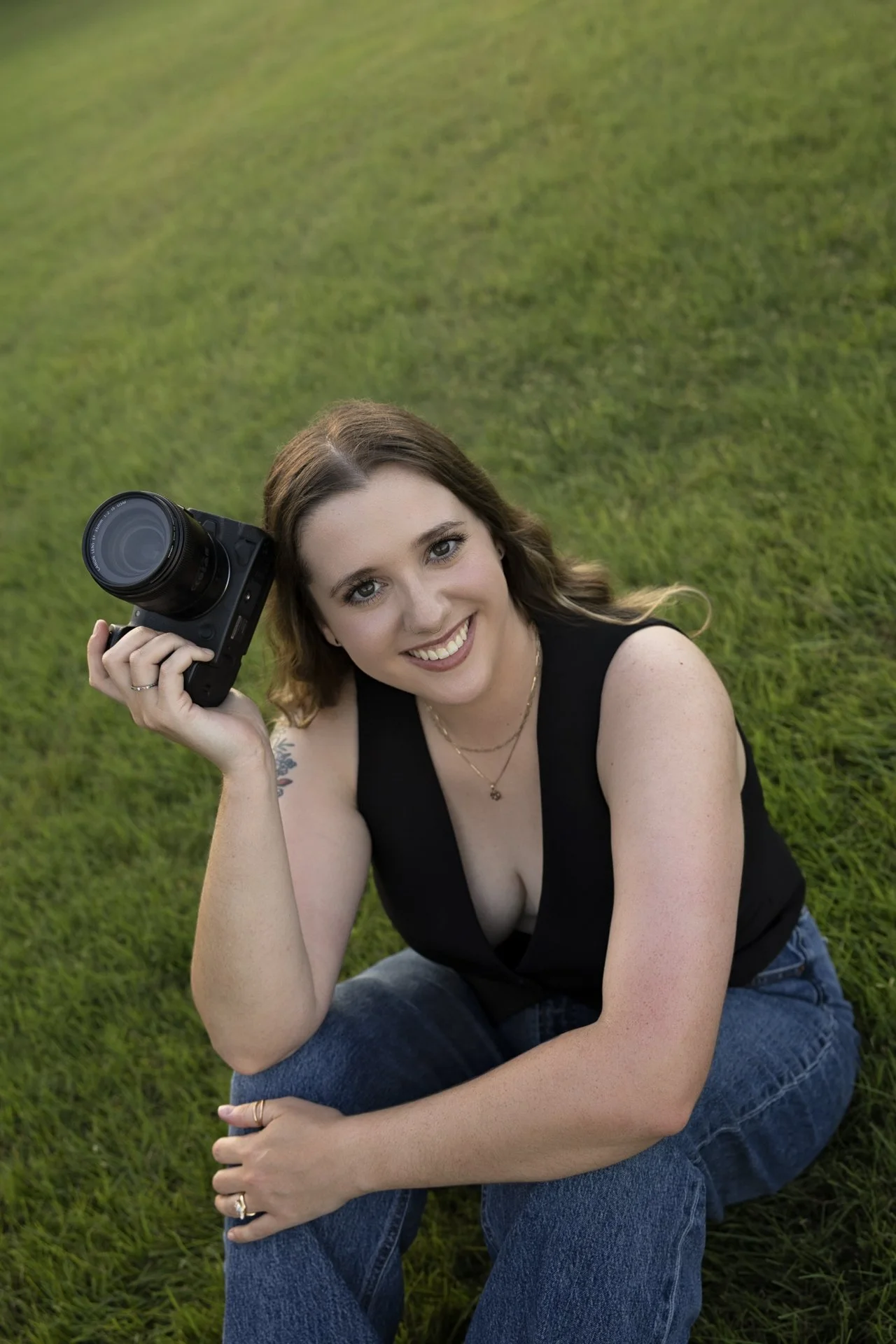 A woman with wavy brown hair, smiling, wearing a black sleeveless top, blue jeans, and jewelry, sitting on grass with a camera in her hand, surrounded by green grass and outdoor natural light.