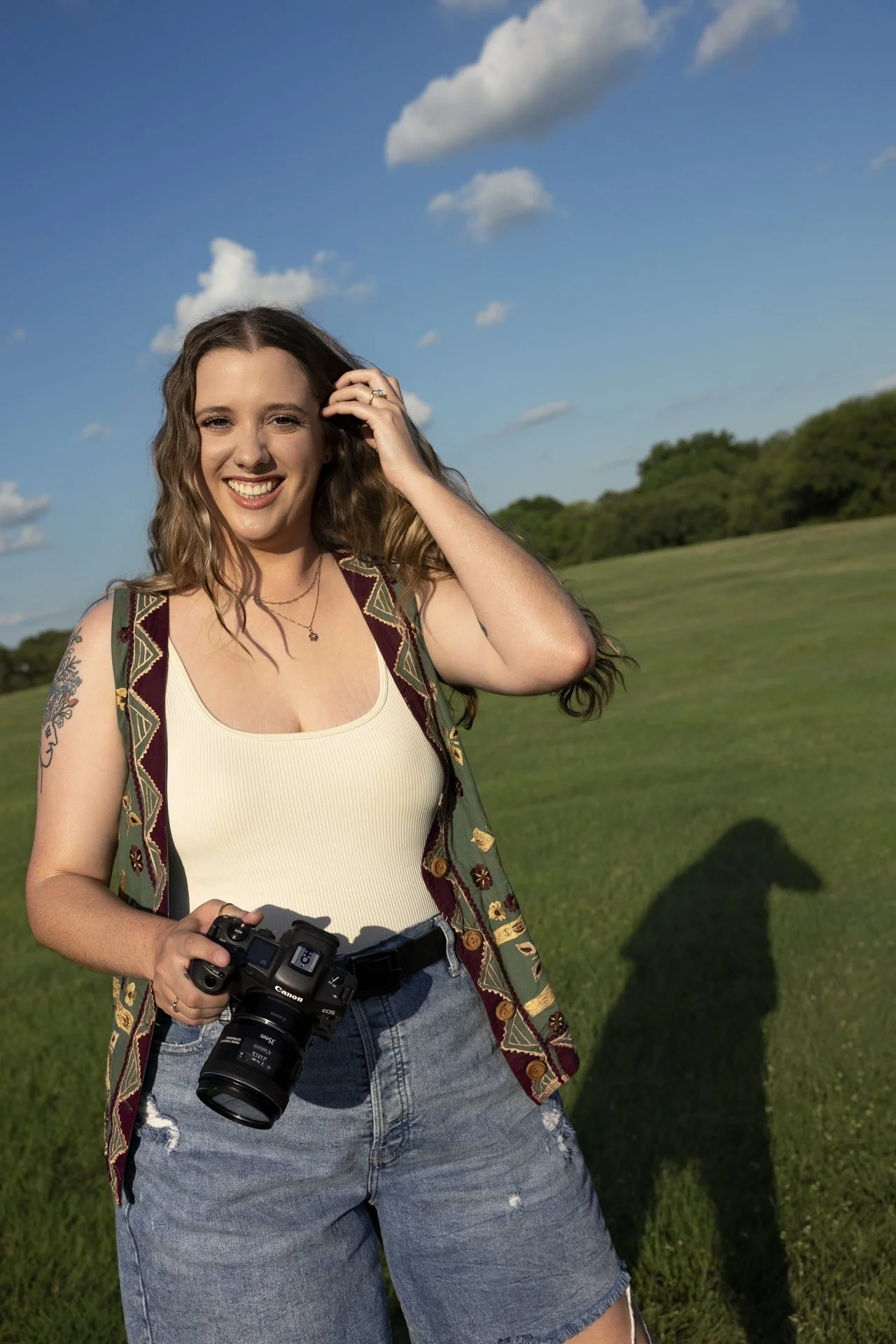 A smiling woman with wavy brown hair holding a camera outdoors in a grassy field with trees in the background and clouds in the sky.