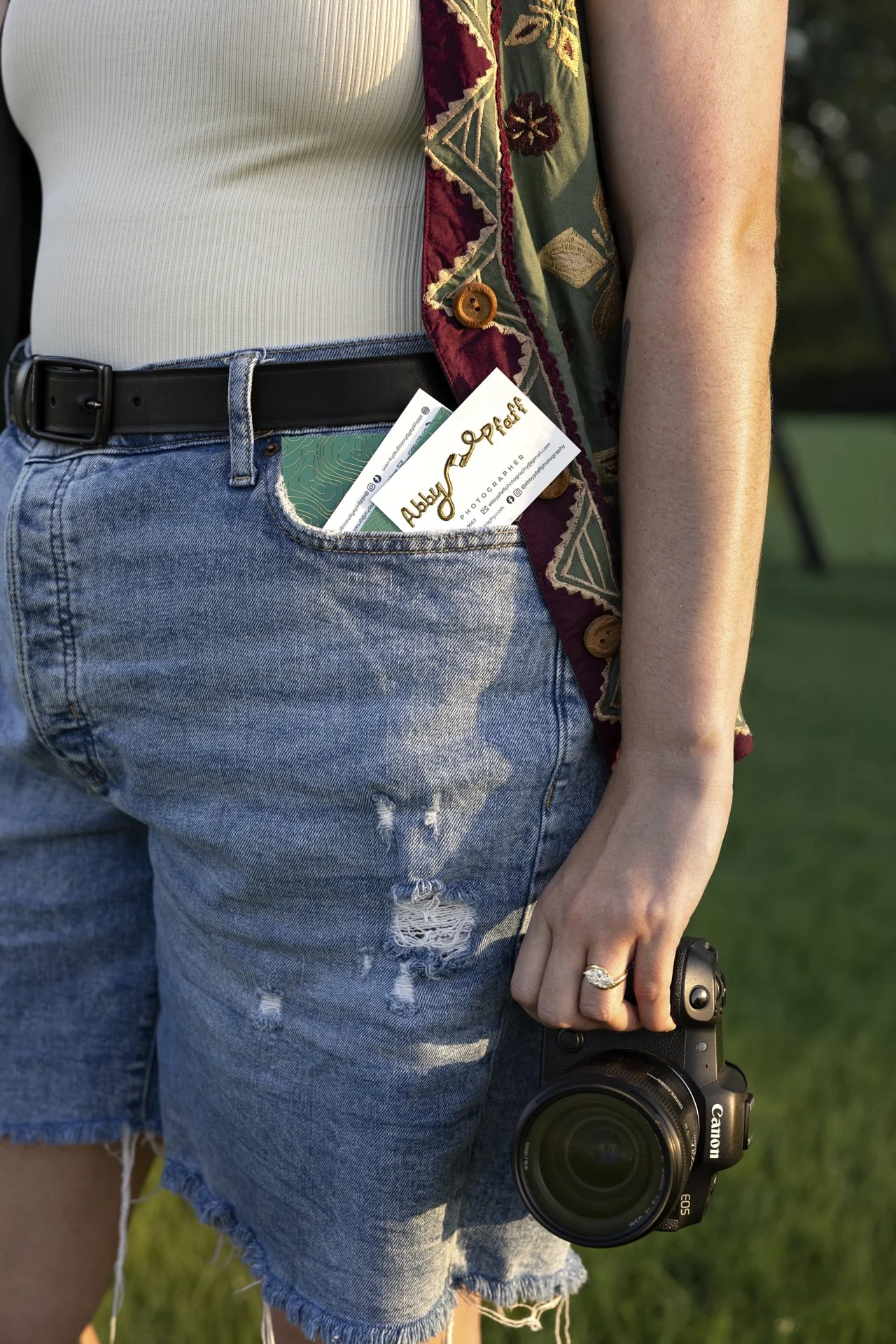 A person wearing a white ribbed tank top, distressed denim shorts, and a colorful embroidered vest, holding a Canon camera in their right hand with a wedding ring on the finger. The person has a business card partially visible in the back pocket of the shorts. The background shows green grass and trees.