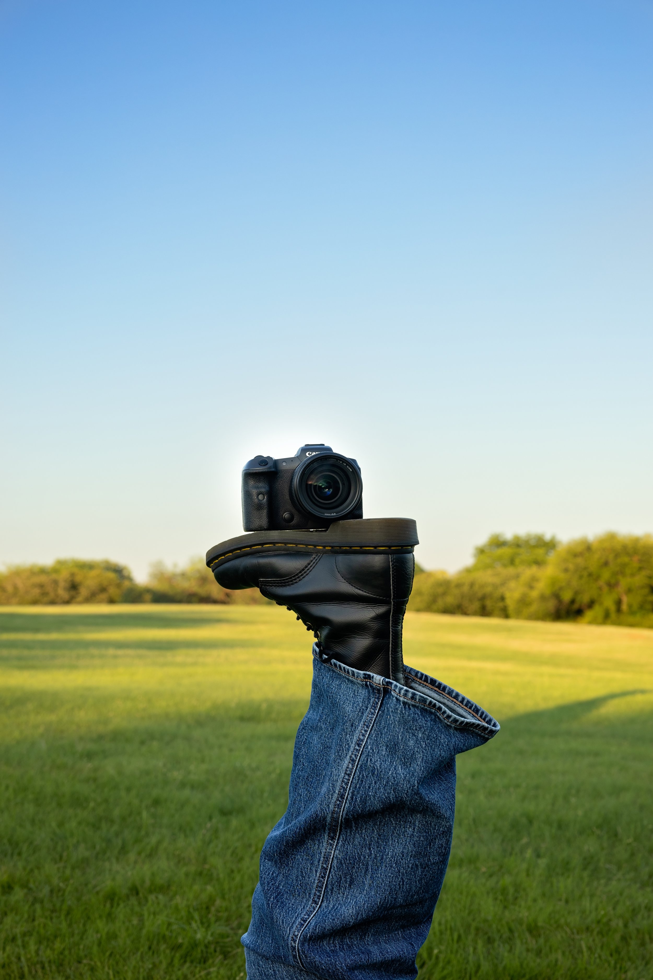 A person wearing jeans and a black glove holding a camera in the sky with a clear blue background, green grassy field and trees in the distance.