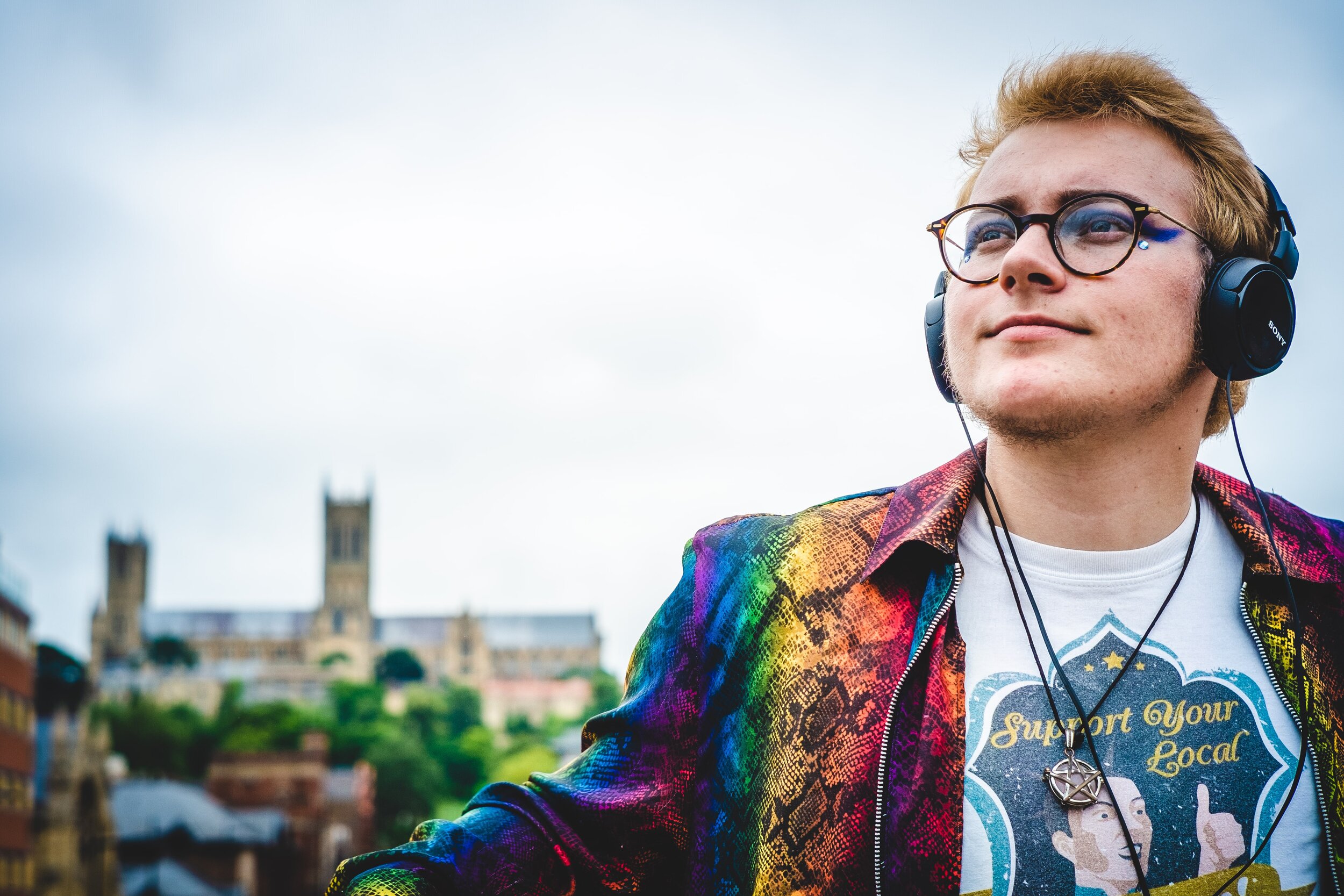 A young person stands, looking into the distance. They wear headphones, and blue eye make up. Behind them out of focus, stands Lincoln Cathedral in distance.