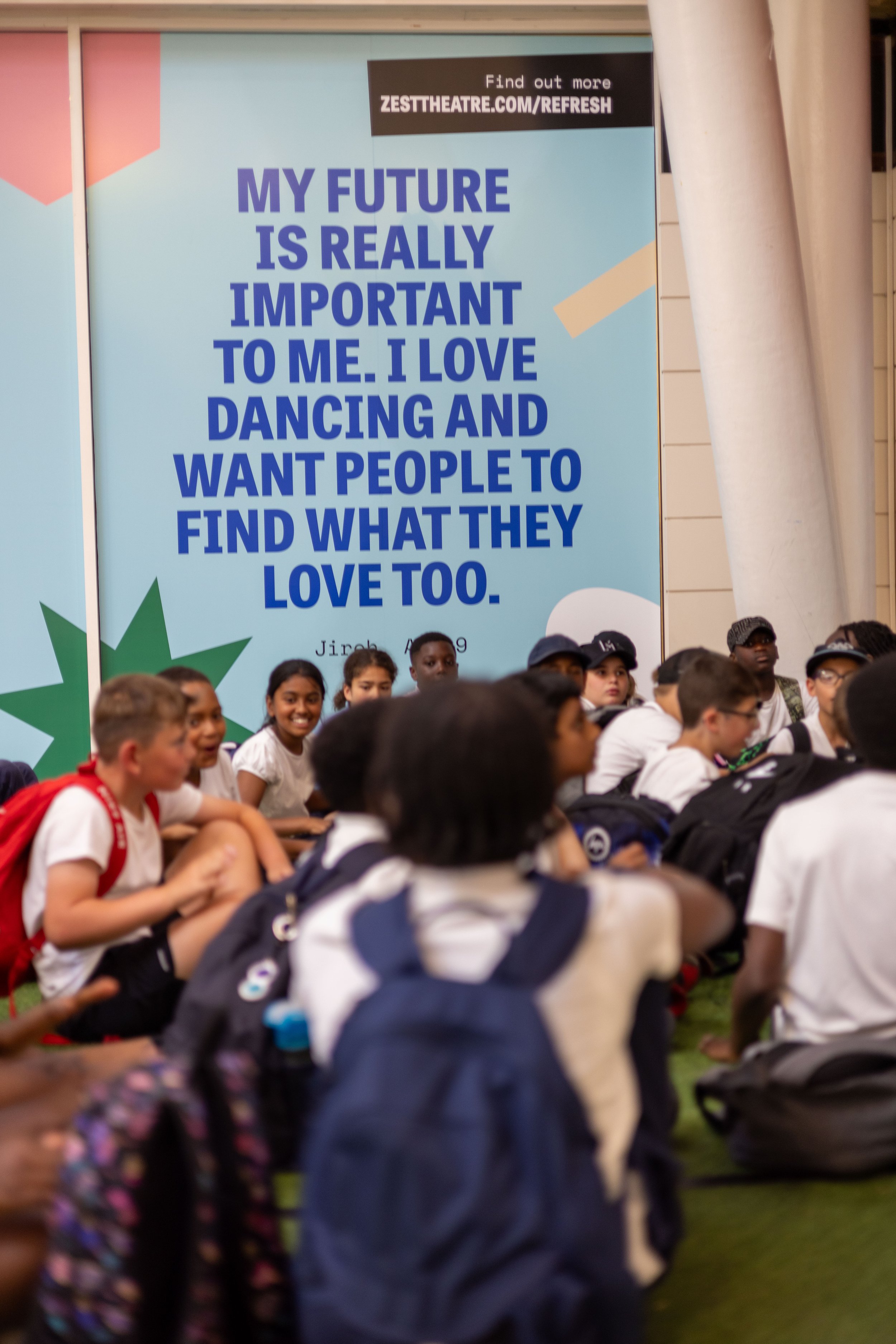A group of kids sit on a shopping centre floor. Behind them colourful artworks with quotes from children loom large.