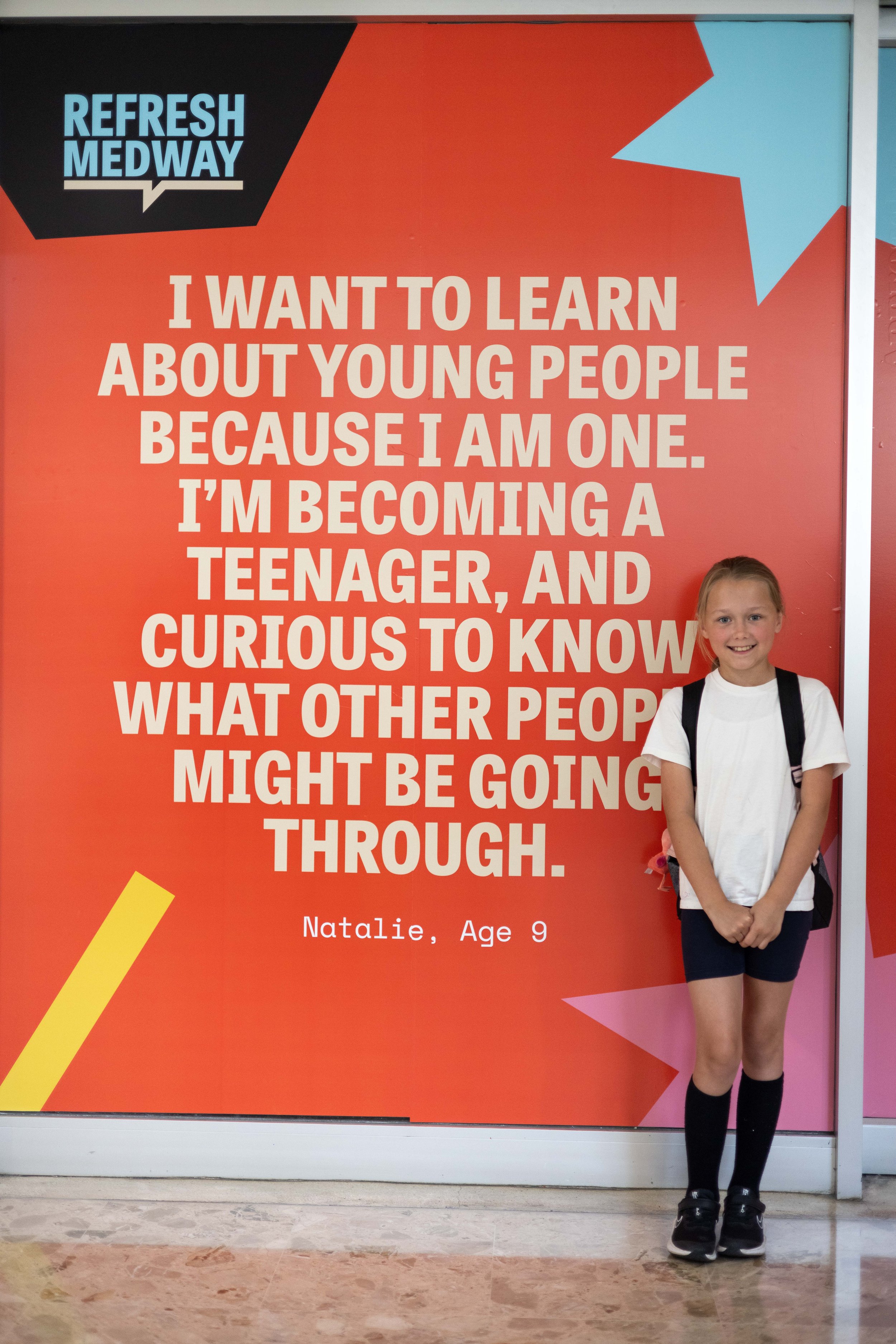 A child smiles to the camera wearing their school uniform. Behind them a colourful artwork wraps an empty shop window. 