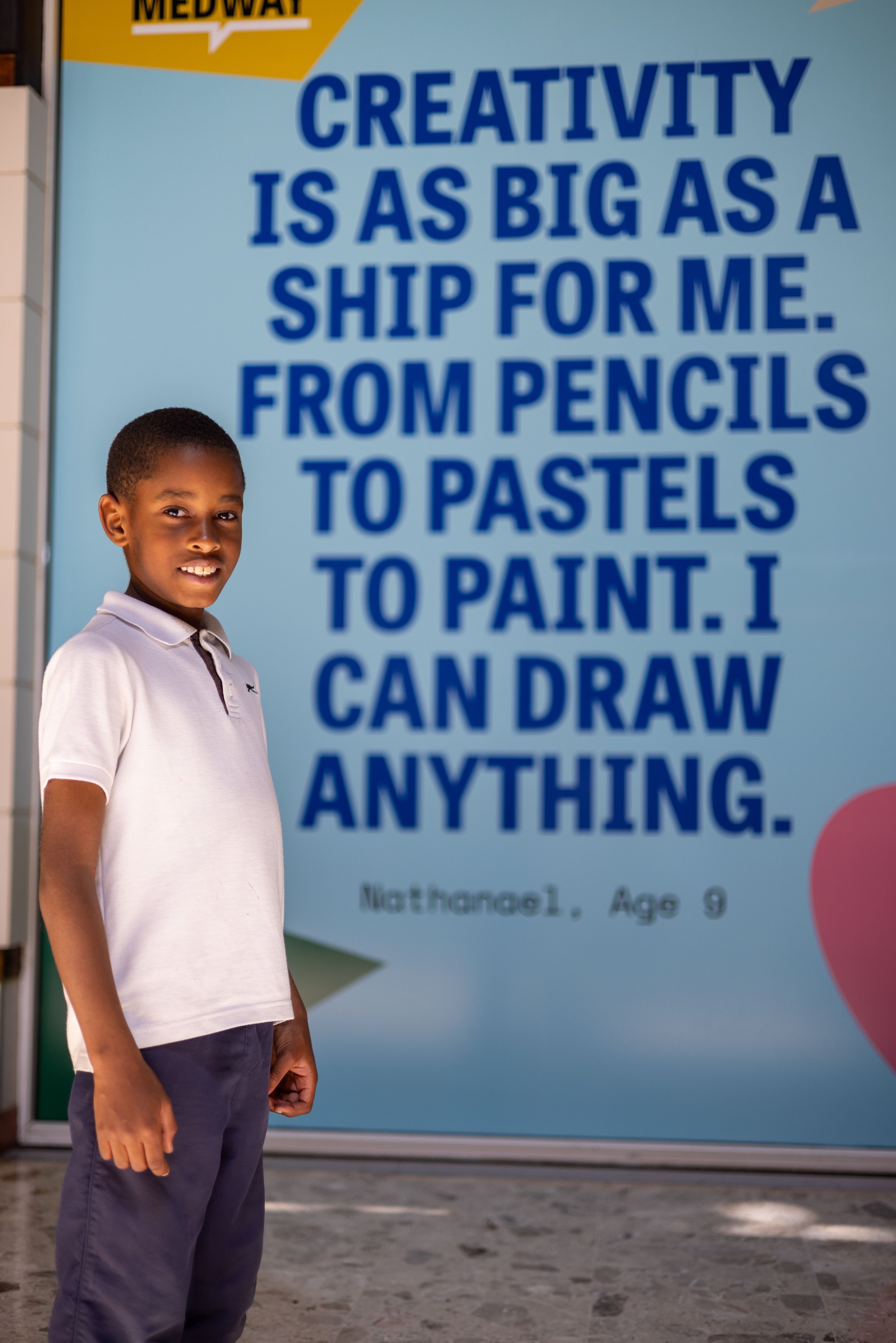 A child smiles to the camera wearing their school uniform. Behind them a colourful artwork wraps an empty shop window. 