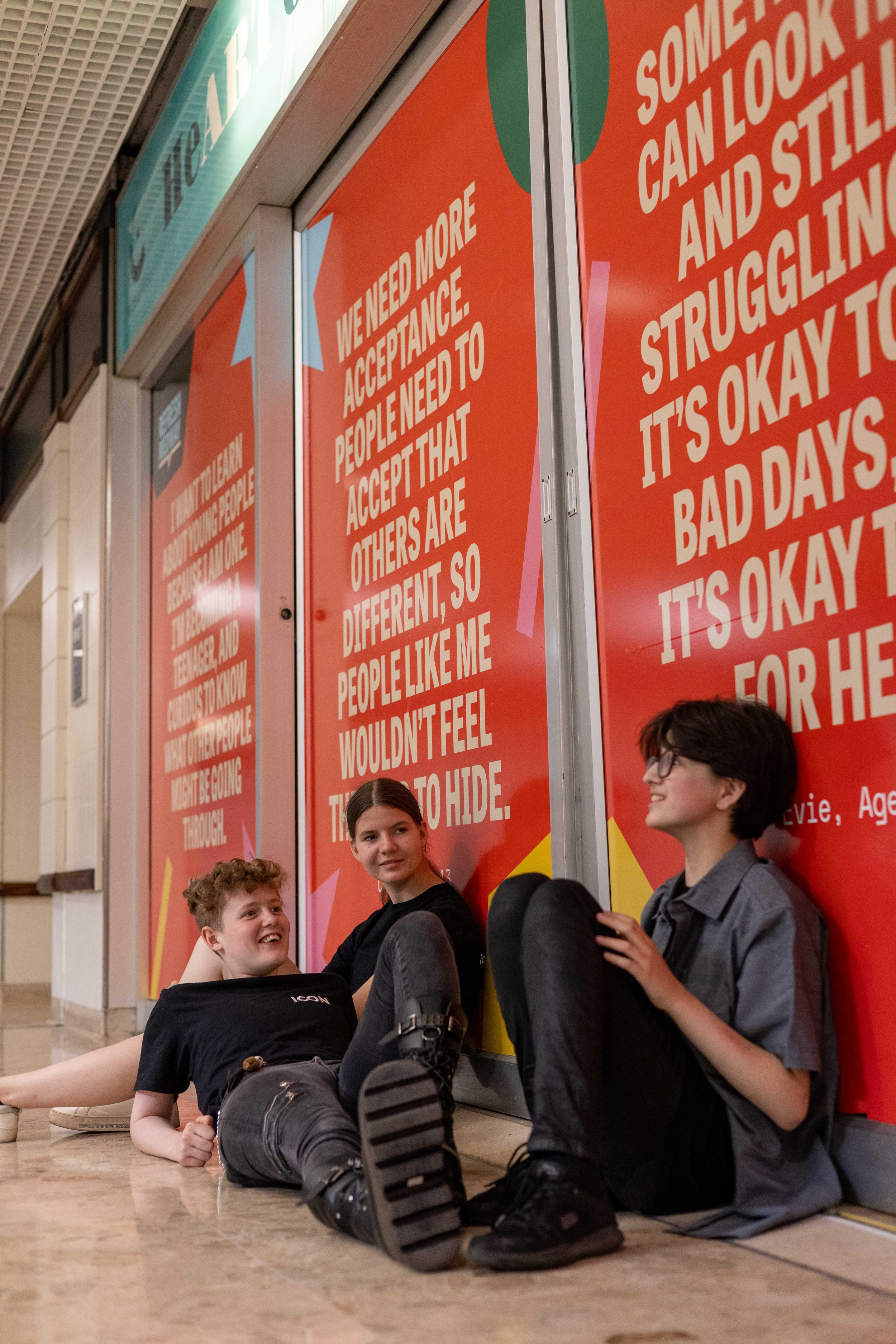 Teenagers hang out on the floor of a shopping centre. They sit in front of a huge artwork featuring the quote of a fellow young person.