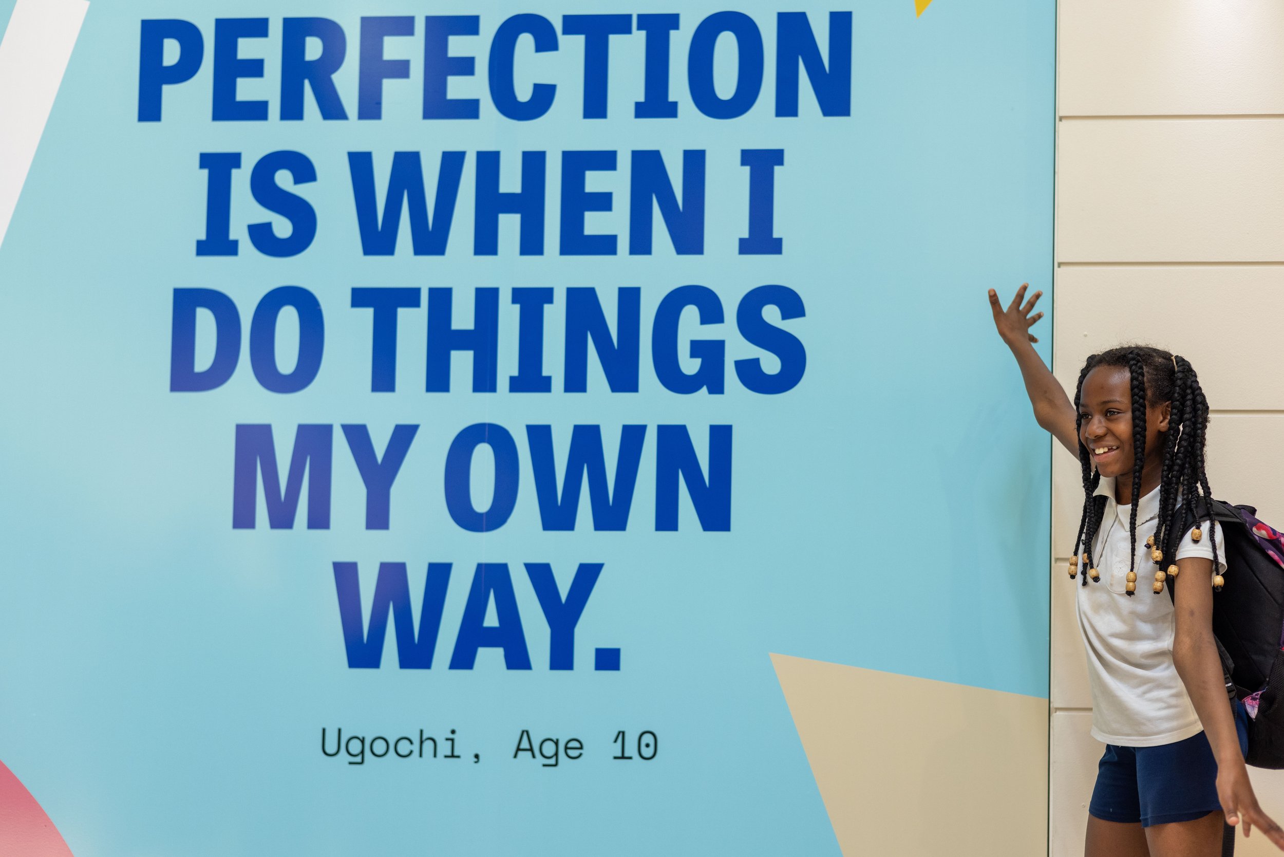 A child stand proudly next to an artwork featuring their quote. 'Perfection is when I do things my own way'.