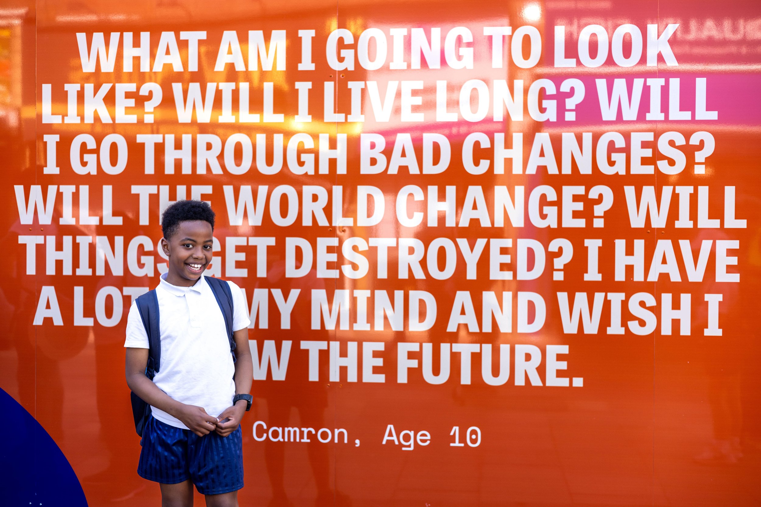 A small child stands in front of an artwork featuring their quote. The quote is bigger than they are. Powerful words from a small human.