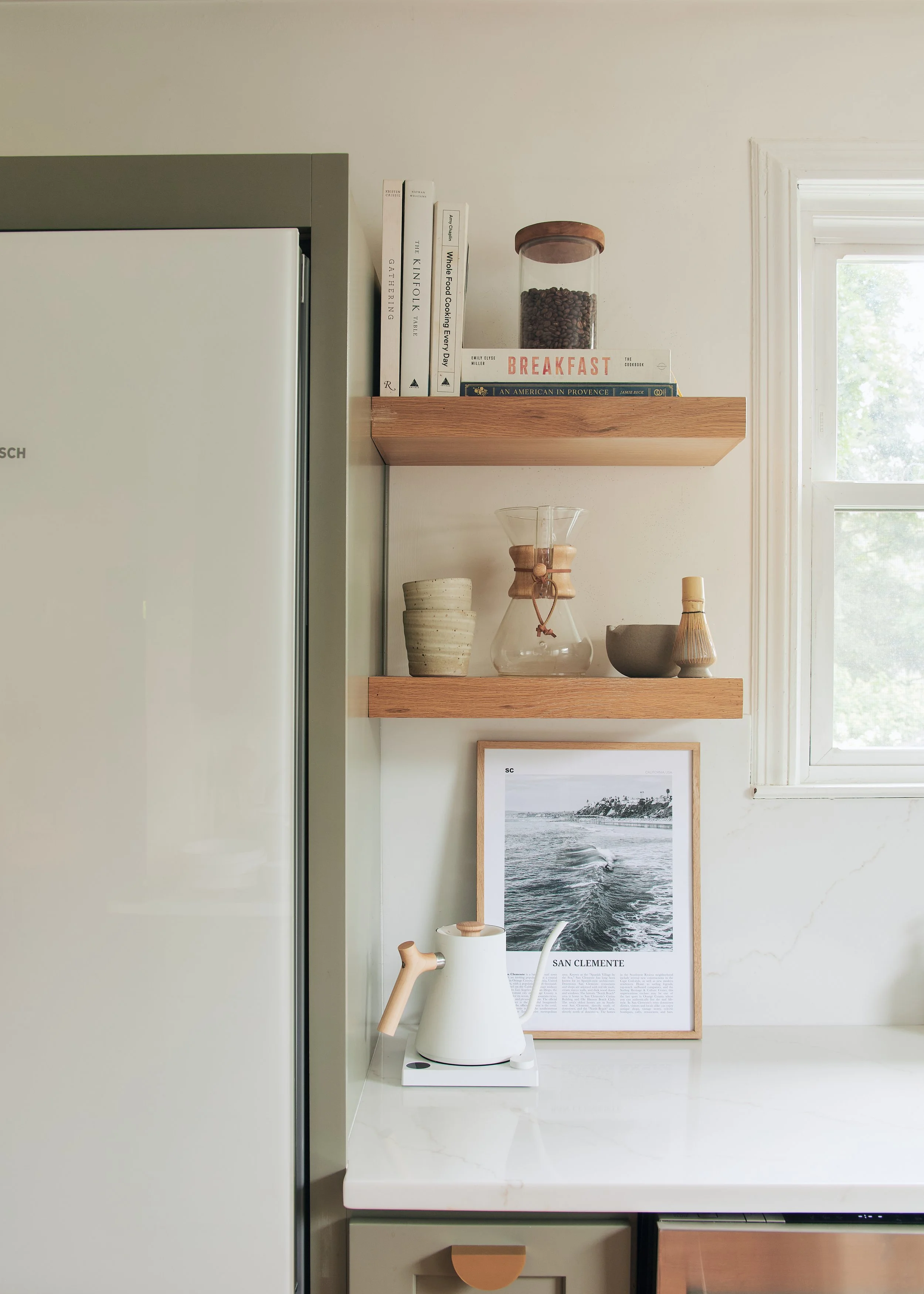 Kitchen with white countertop, a white kettle, floating wooden shelves holding books, ceramics, a glass coffee pot, and a framed black-and-white photograph near a window.
