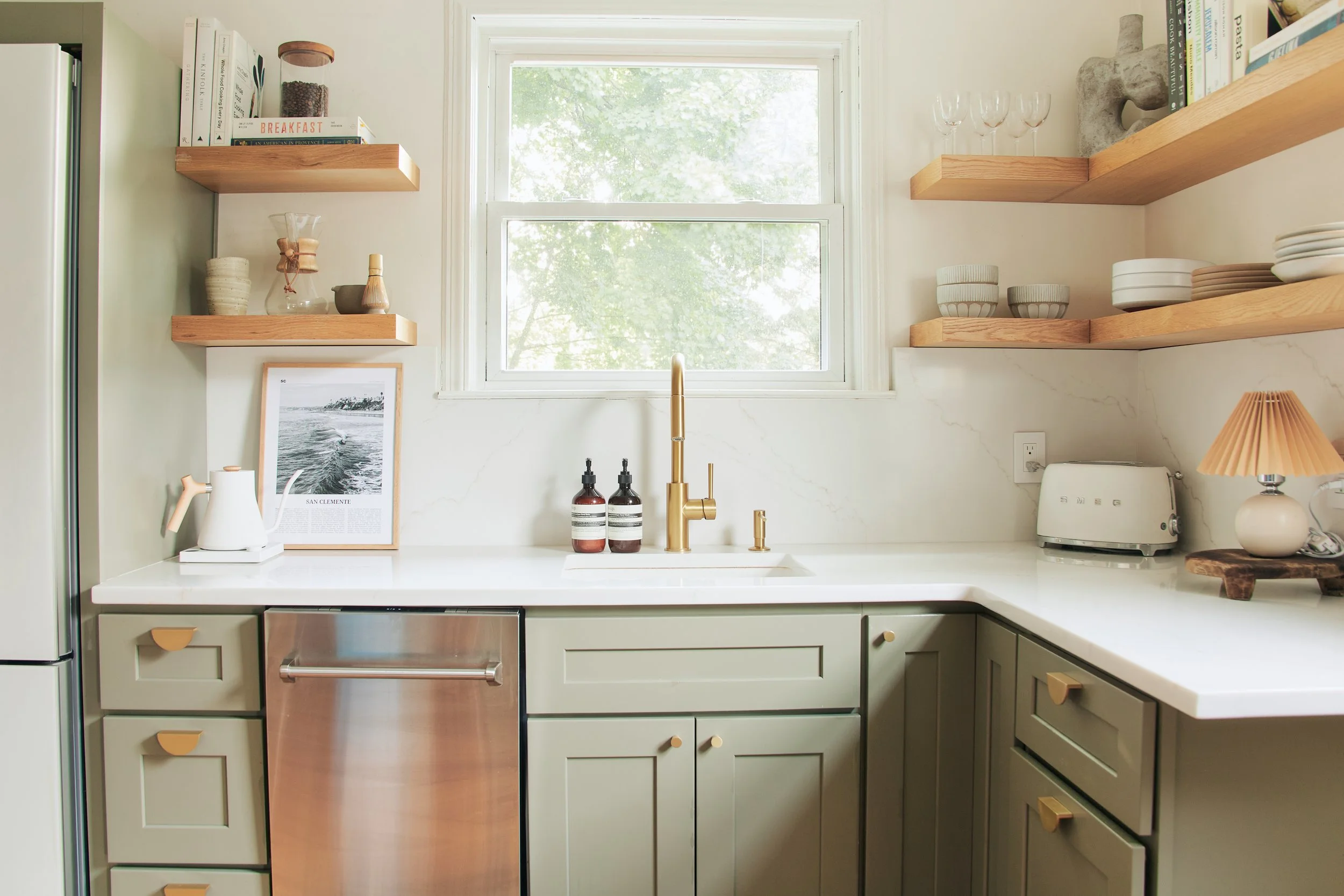 Clean modern kitchen with green cabinets, white countertop, and gold faucet. Open wooden shelves hold dishes and glassware. Small kitchen appliances include a toaster and a lamp with a pleated shade.