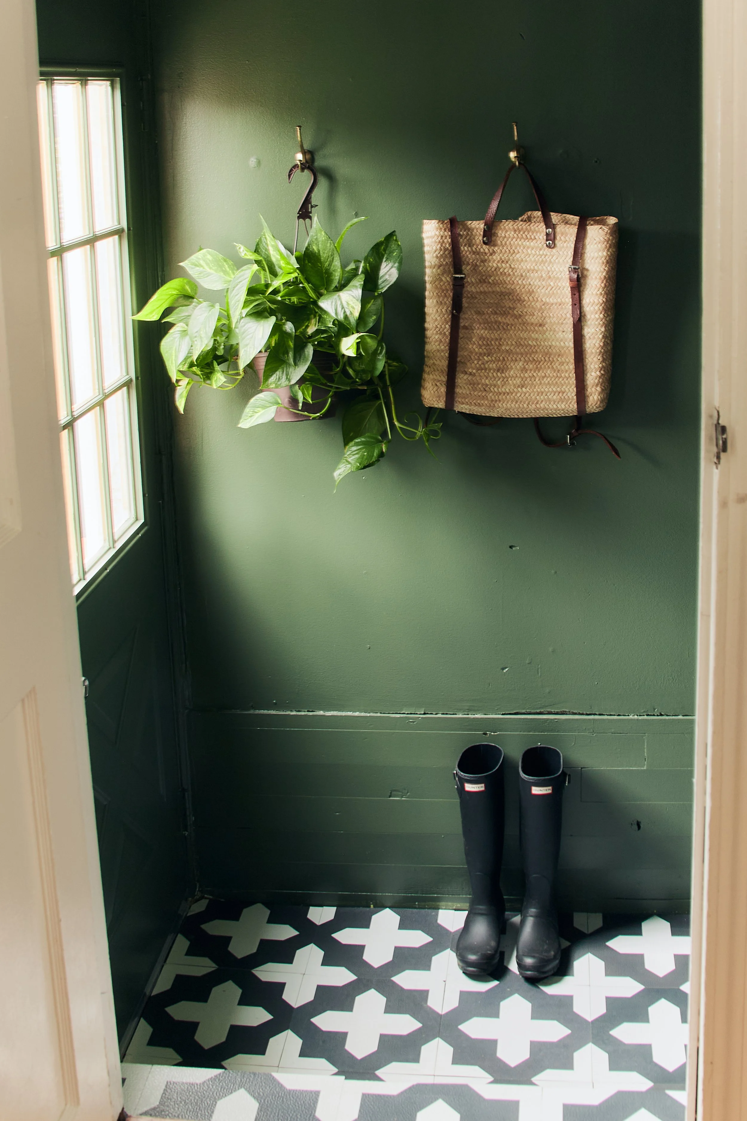 A small entryway with green walls, a window on the left, a hanging green plant, a woven tote bag, and a pair of black rain boots on a patterned black and white tile floor.