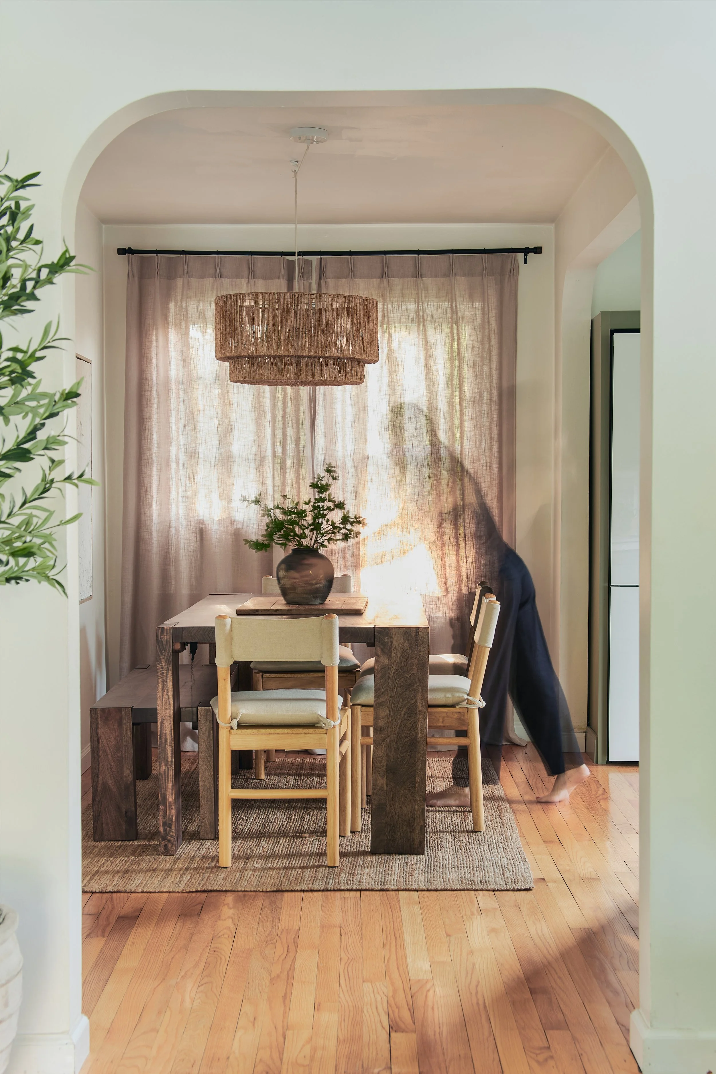 Dining area with wooden table, beige chairs, a potted plant on the table, pink curtains, and a woven pendant light fixture.