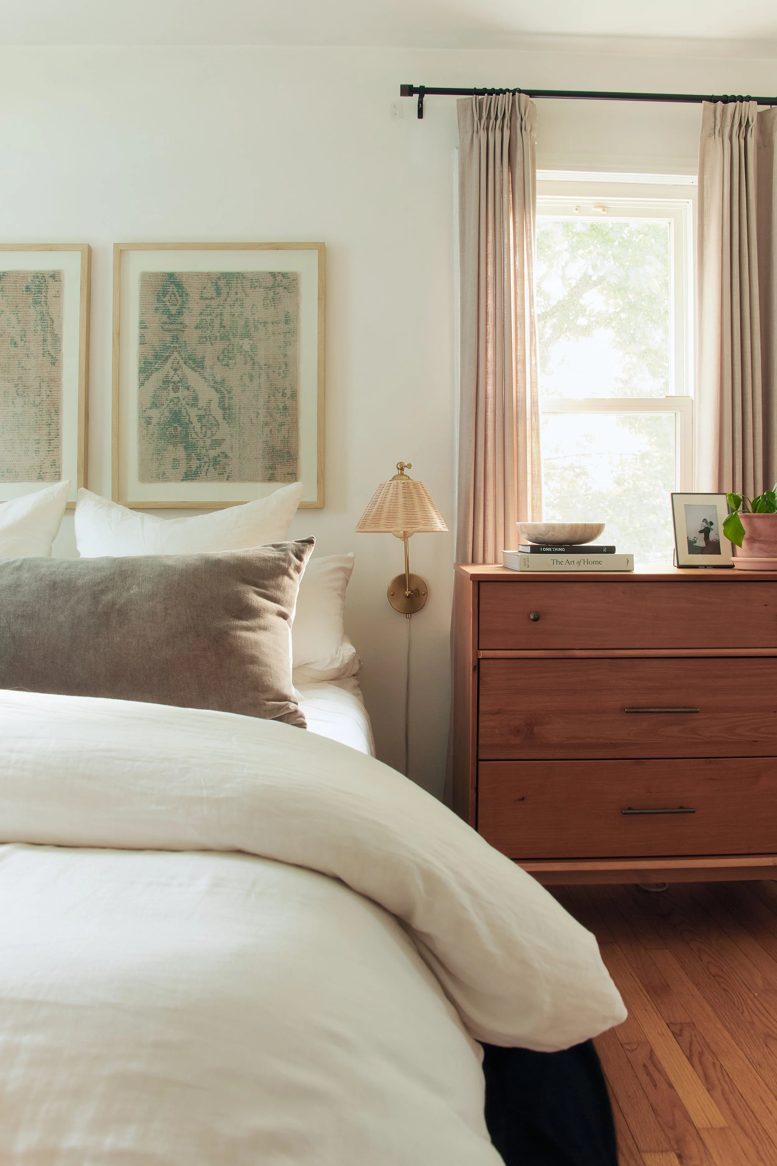 A cozy bedroom corner featuring a bed with white and beige bedding, decorative pillows, framed artwork on the wall, a wooden dresser with books, a bowl, and a framed photo, and a window with light pink curtains.