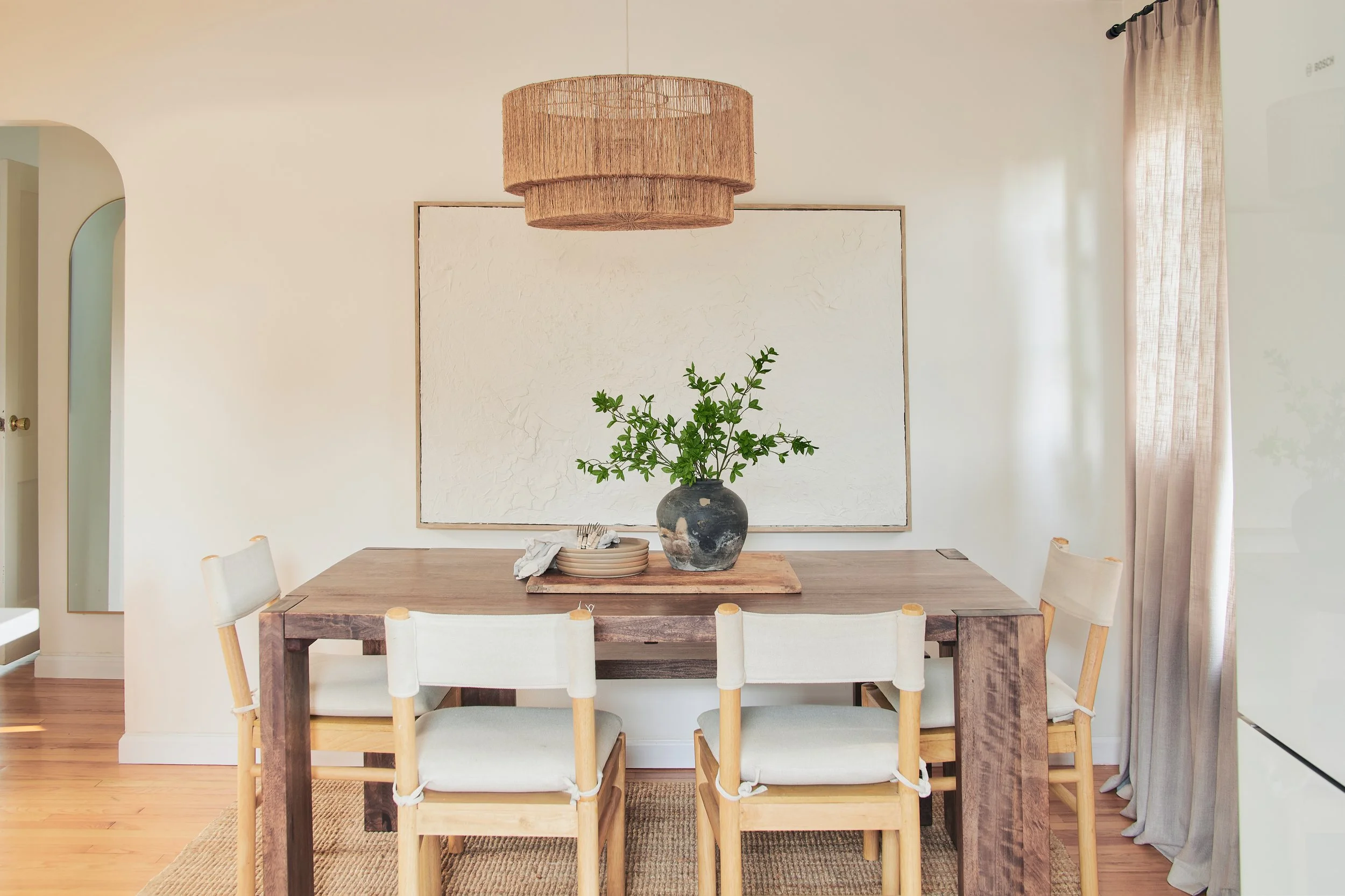 A wooden dining table with four chairs, a vase with green foliage, and a place setting, in a bright dining room with beige walls, a textured wall art piece, and a window with light-colored curtains.