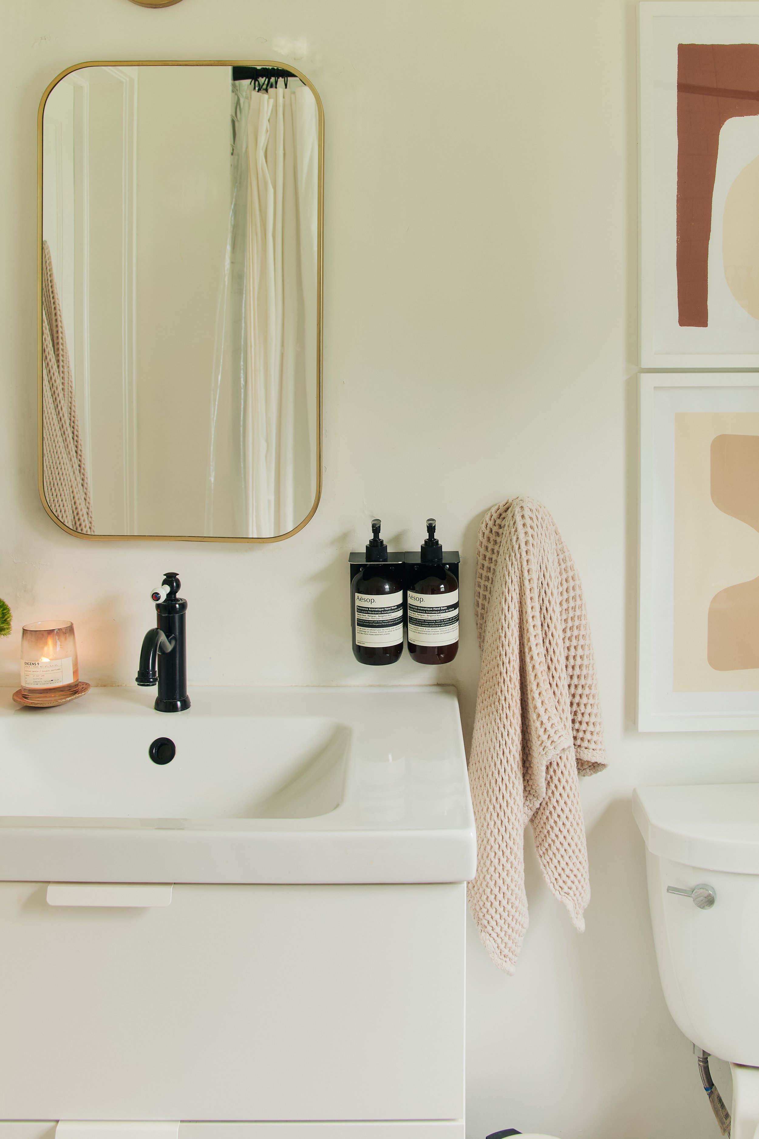 Bathroom vanity with a mirror, a candle, a black faucet, two wall-mounted bottles, a beige towel hanging on a hook, and part of a white toilet.