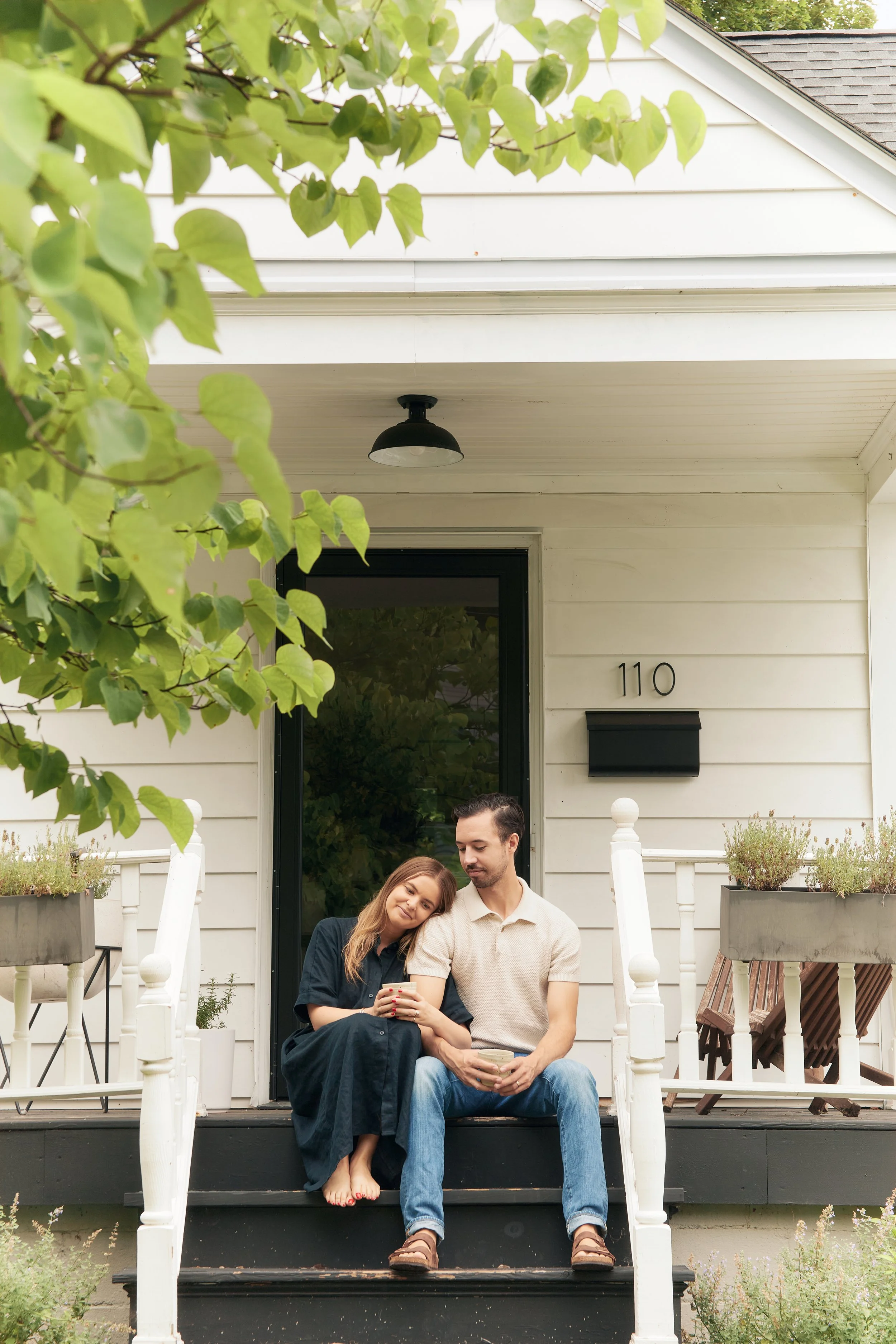 A couple sitting on the front porch of a house with white siding, surrounded by plants and trees, smiling and enjoying each other's company.