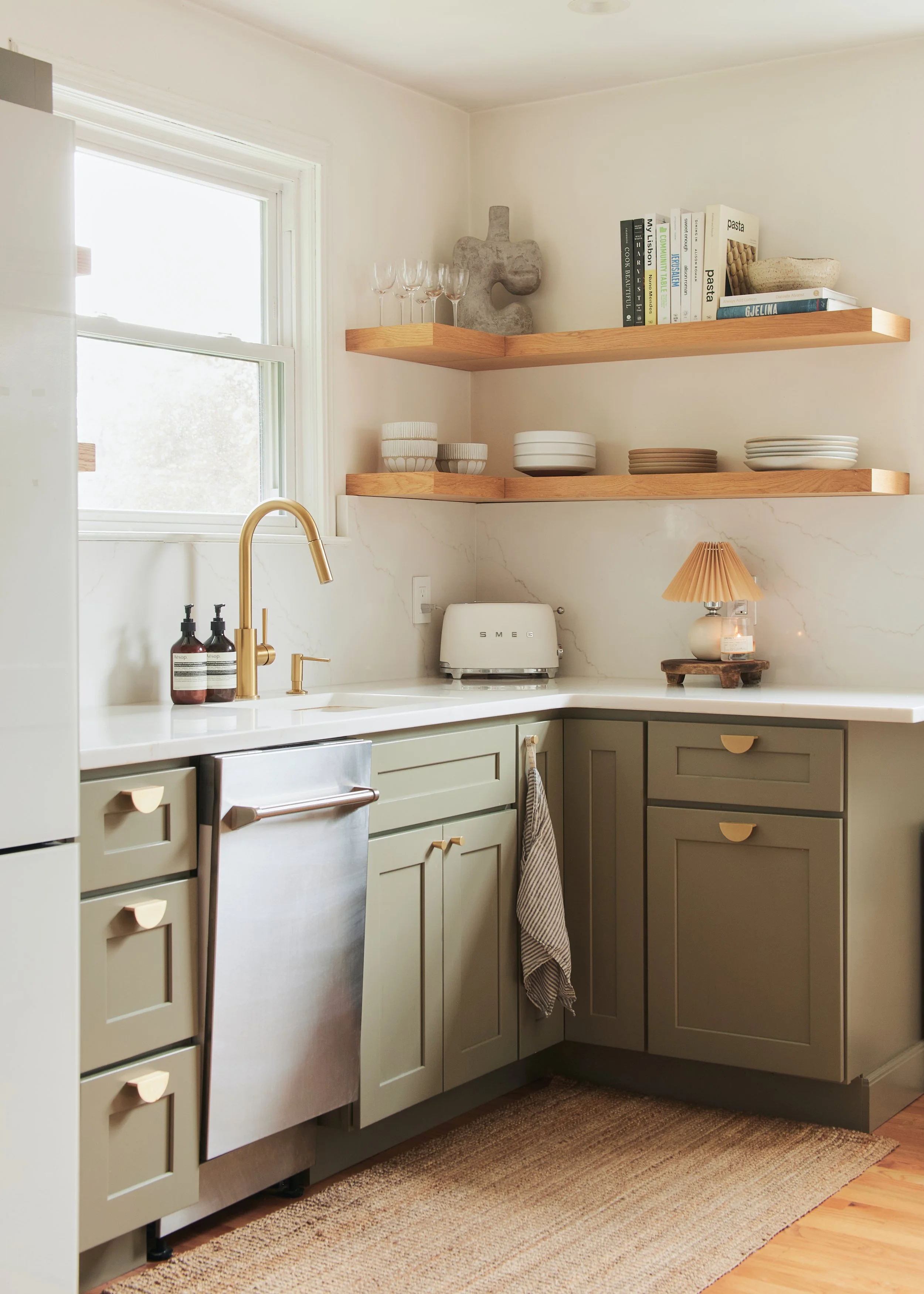 Kitchen with light green cabinetry, white marble countertops, gold faucet, open shelves with dishes and books, window, small lamp, and toaster.