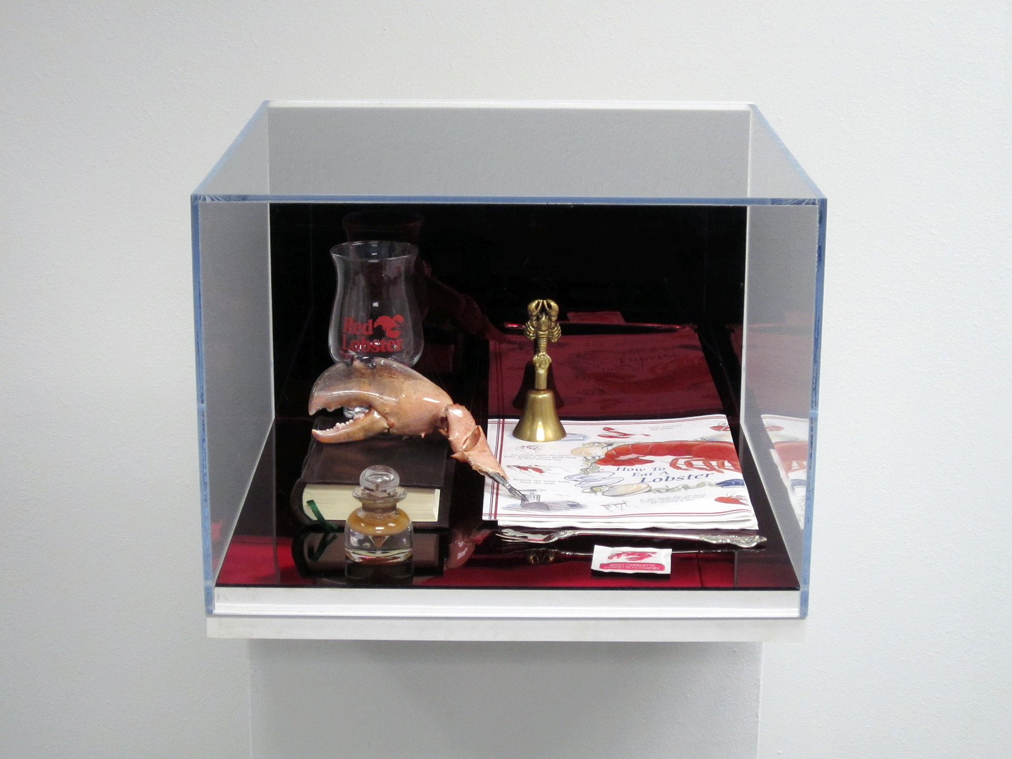   How to Eat Lobster (Ritual)  2011 Wall mounted plexi-glass and wood vitrines, contents: red mirrored plexi-glass, "Red Lobster" drinking glass, brass bell, paper placemat and napkin, leather book, moist towelette, sterling silver lobster fork, taxi