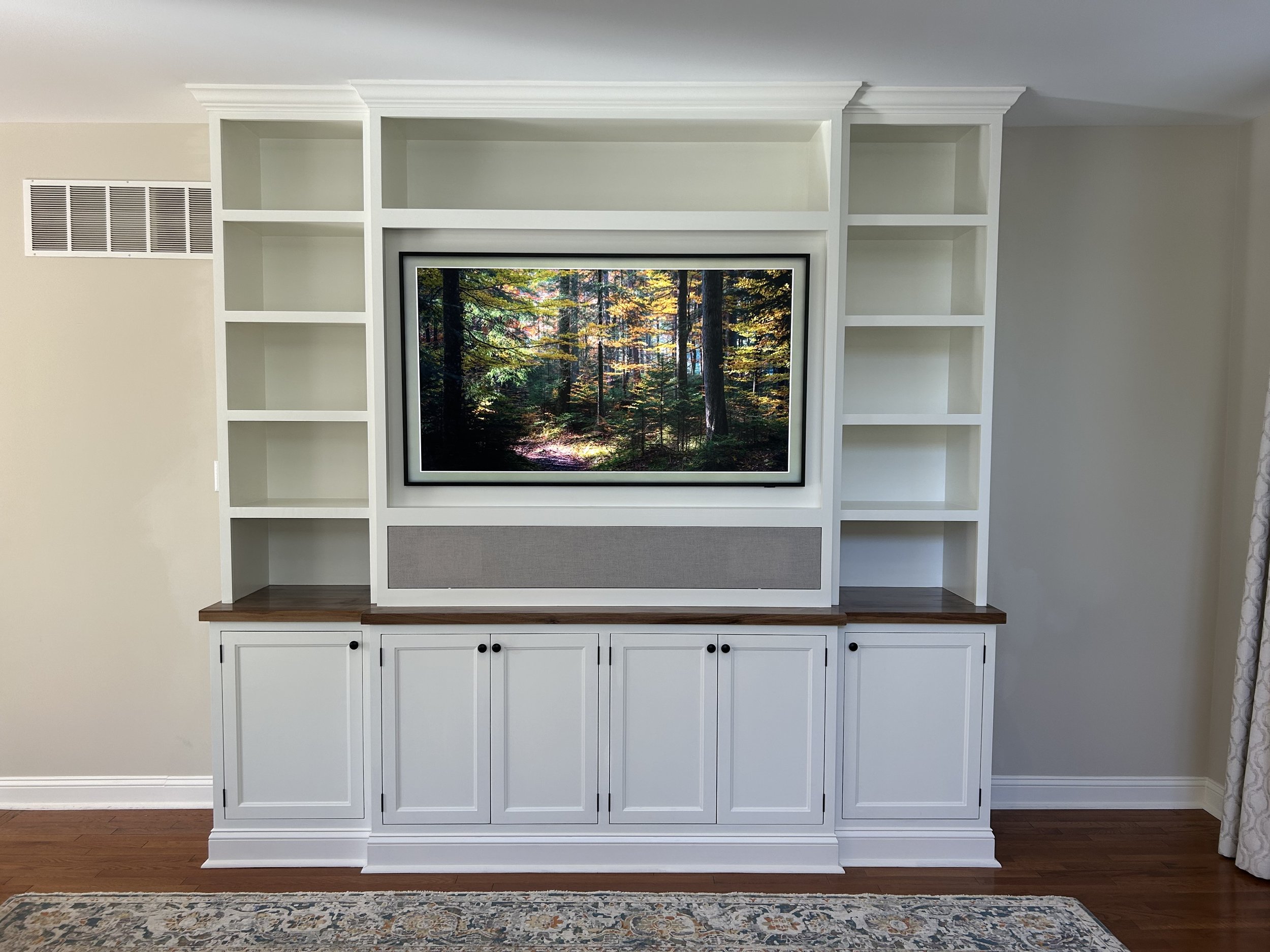  This living entertainment center features inset doors, a black walnut countertop, and upper shelving surrounding a central frame TV. Below the TV is an open area which houses the audio equipment and speakers, which are hidden behind a fabric covered