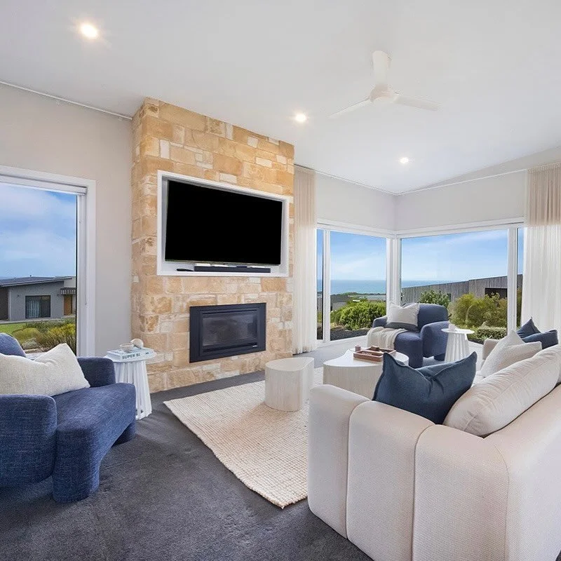 This living room steals the show! Sweeping ocean views, a warm sandstone fireplace, and a palette that mirrors the calm of the sea itself.

We styled it with soft, muted blue tones and natural textures. Think travertine coffee tables, layered linen, 