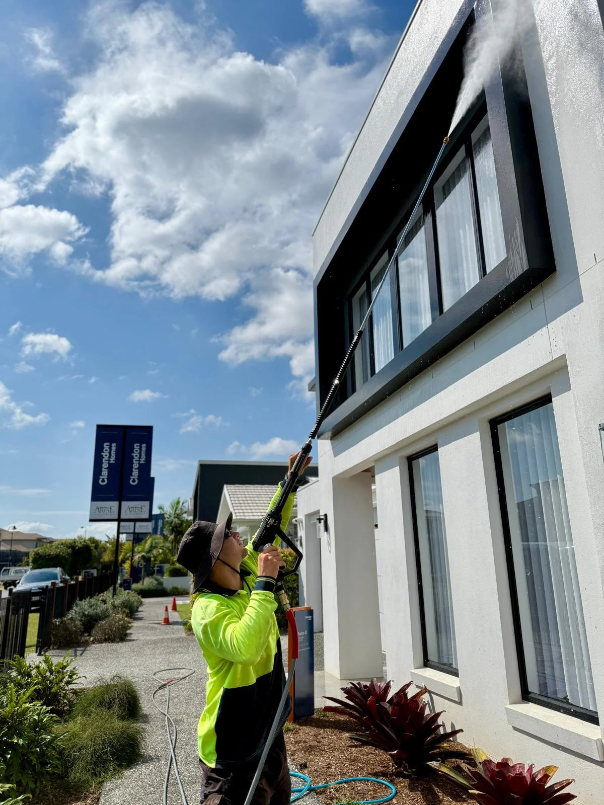 A person in a bright yellow jacket and hat pressure washing the exterior of a white modern building with large windows on a sunny day.