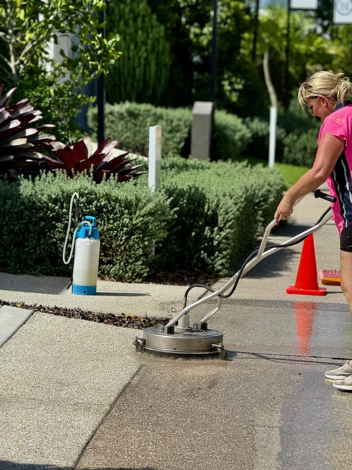 Woman using a sidewalk cleaning machine to wash the pavement outdoors in a park-like area with bushes and trees.