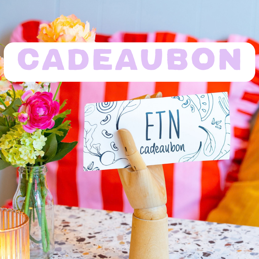 Een geschenkbon op een houten standaard met bloemen en kaars op een tafel. Maak iemand blij met een cadeaubon van ETN. 