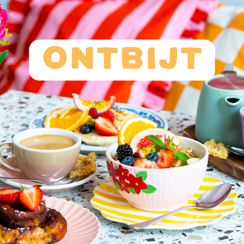 Een ontbijttafel met een kom met fruit, een kop koffie, een pannenkoek met fruit, en een stuk brood met chocolade en aardbeien, op een tafel met kleurrijke achtergrond. Ontbijten bij ETN in Enschede.