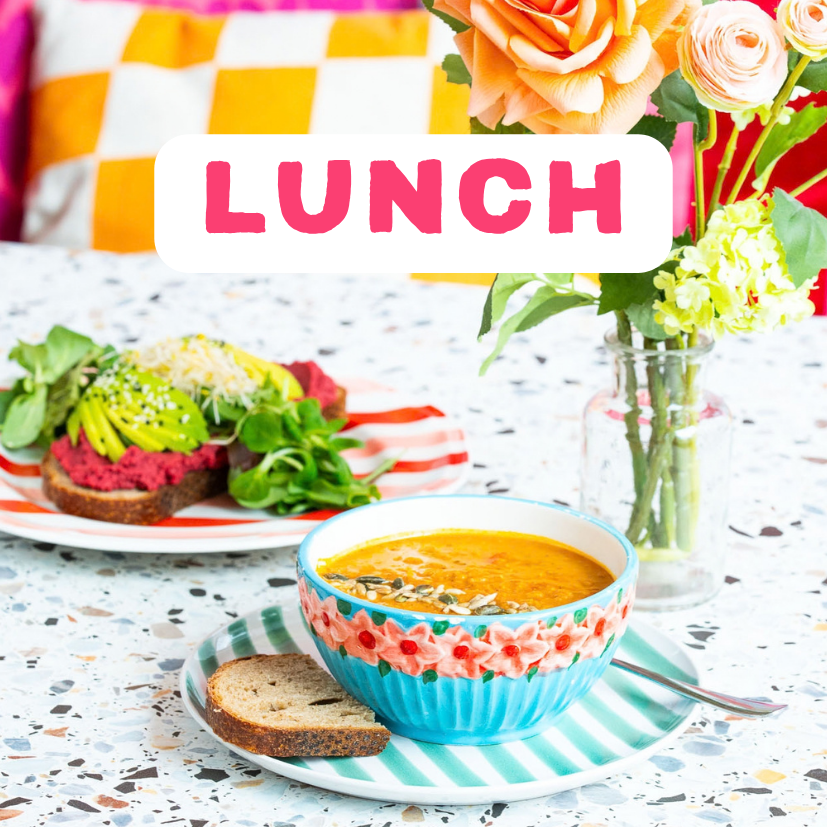 Een kleurrijke tafel met een kom soep, een stukje brood, een avocadotoast met rucola en biet, en een vaas met bloemen, met overlaytekst 'LUNCH' in roze letters. Lunchen bij ETN in Enschede.