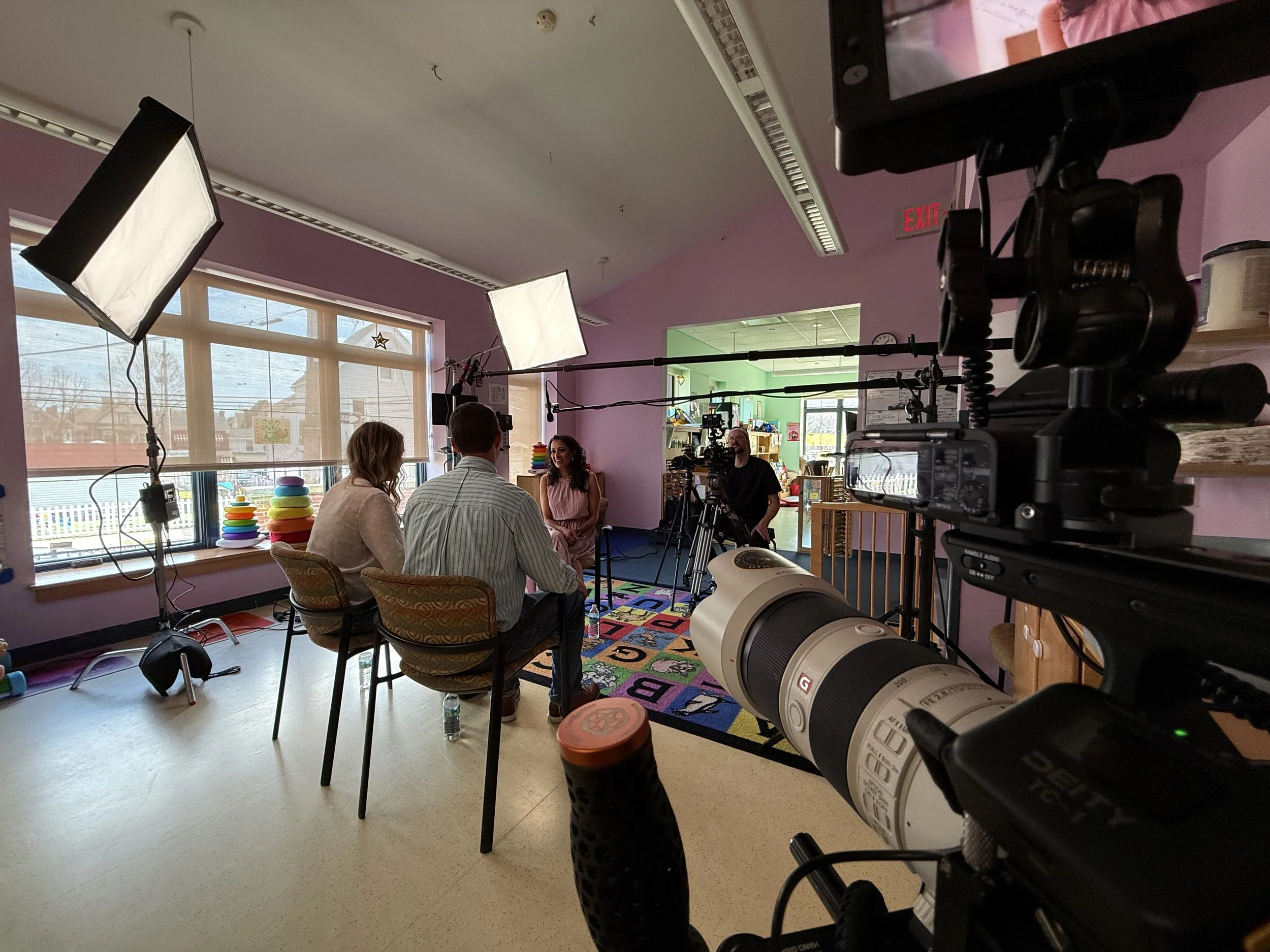 Behind the scenes of a video or interview shoot in a brightly lit room with pink walls, featuring a woman smiling and talking, two people seated and engaged, professional camera and lighting equipment, large windows, colorful stacking rings, and an a