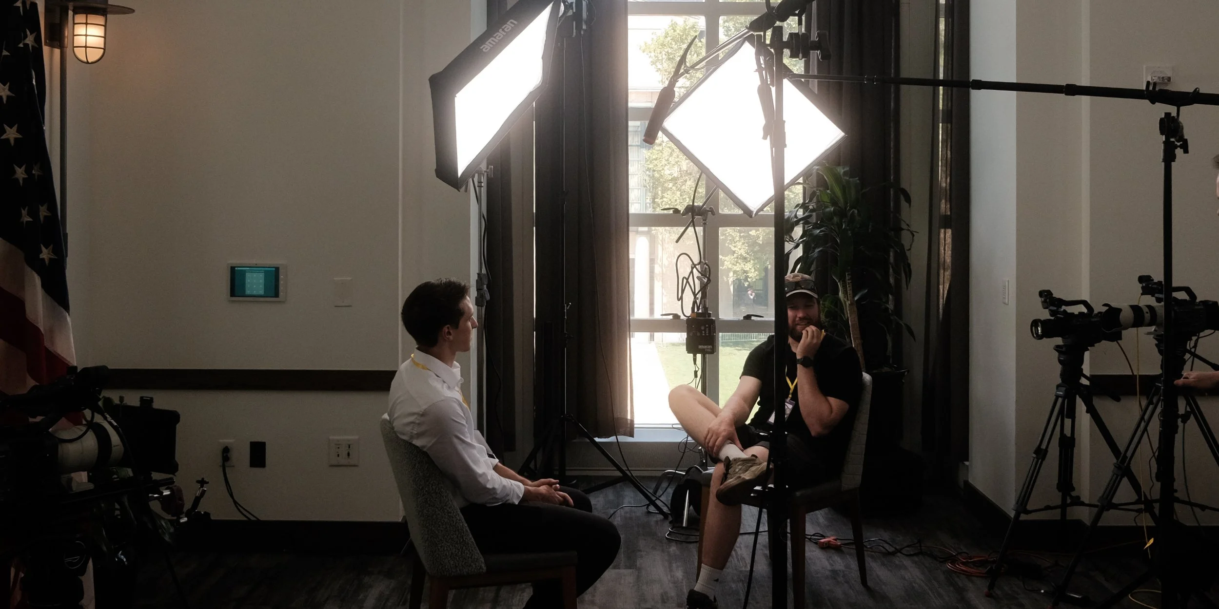 A video interview setup with two men sitting in chairs facing each other. The man on the left is wearing a white shirt, and the man on the right is wearing a black shirt, baseball cap, and shorts. They are surrounded by professional lighting equipmen