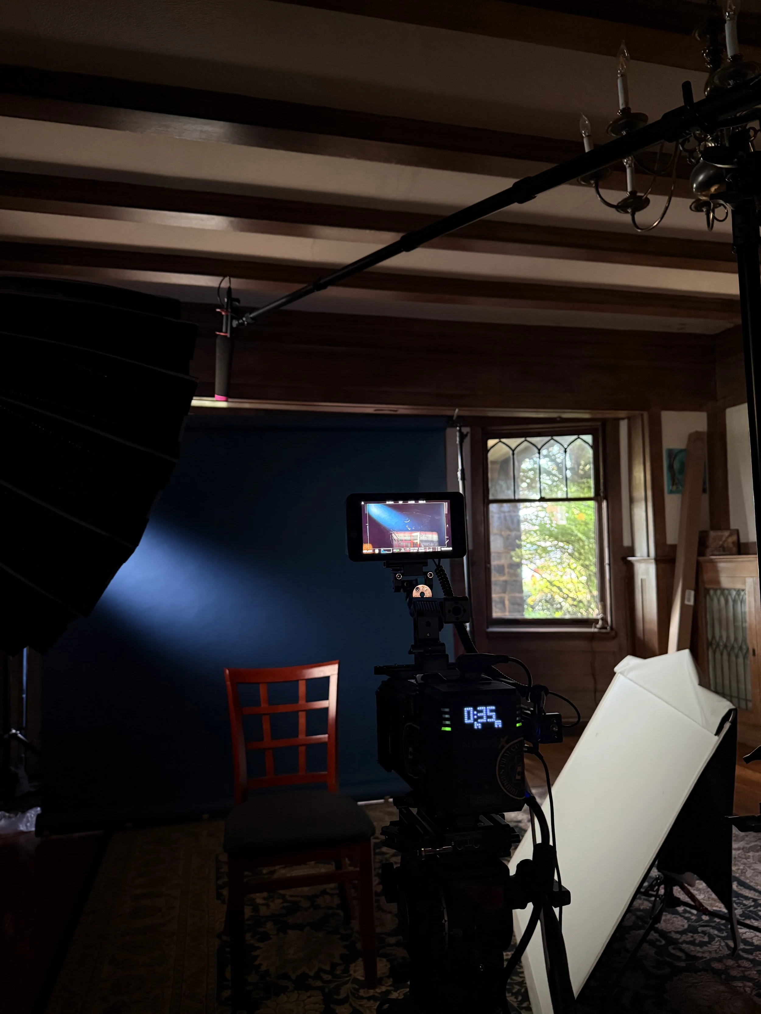 A film or photography set within a room featuring a blue backdrop, a bright light source, a red chair, and filming equipment including a monitor displaying a scene, a camera, and lighting tools. Large windows with daylight, wooden ceiling beams, and 